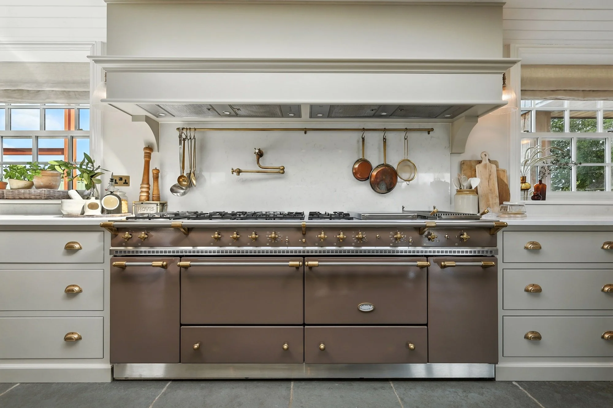 A modern kitchen with a large silver stove with brass handles and knobs, white cabinets, a marble countertop, and a backsplash with hanging copper pots and utensils. There are windows and some potted plants in the background.