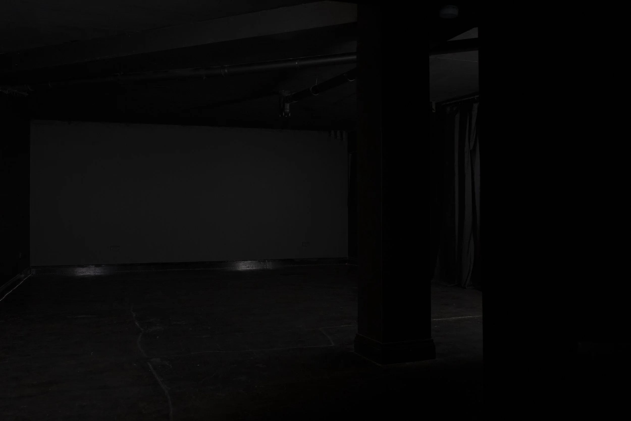 A dark room with faint lighting, a curtain on the right, and some visible pipes on the ceiling.
