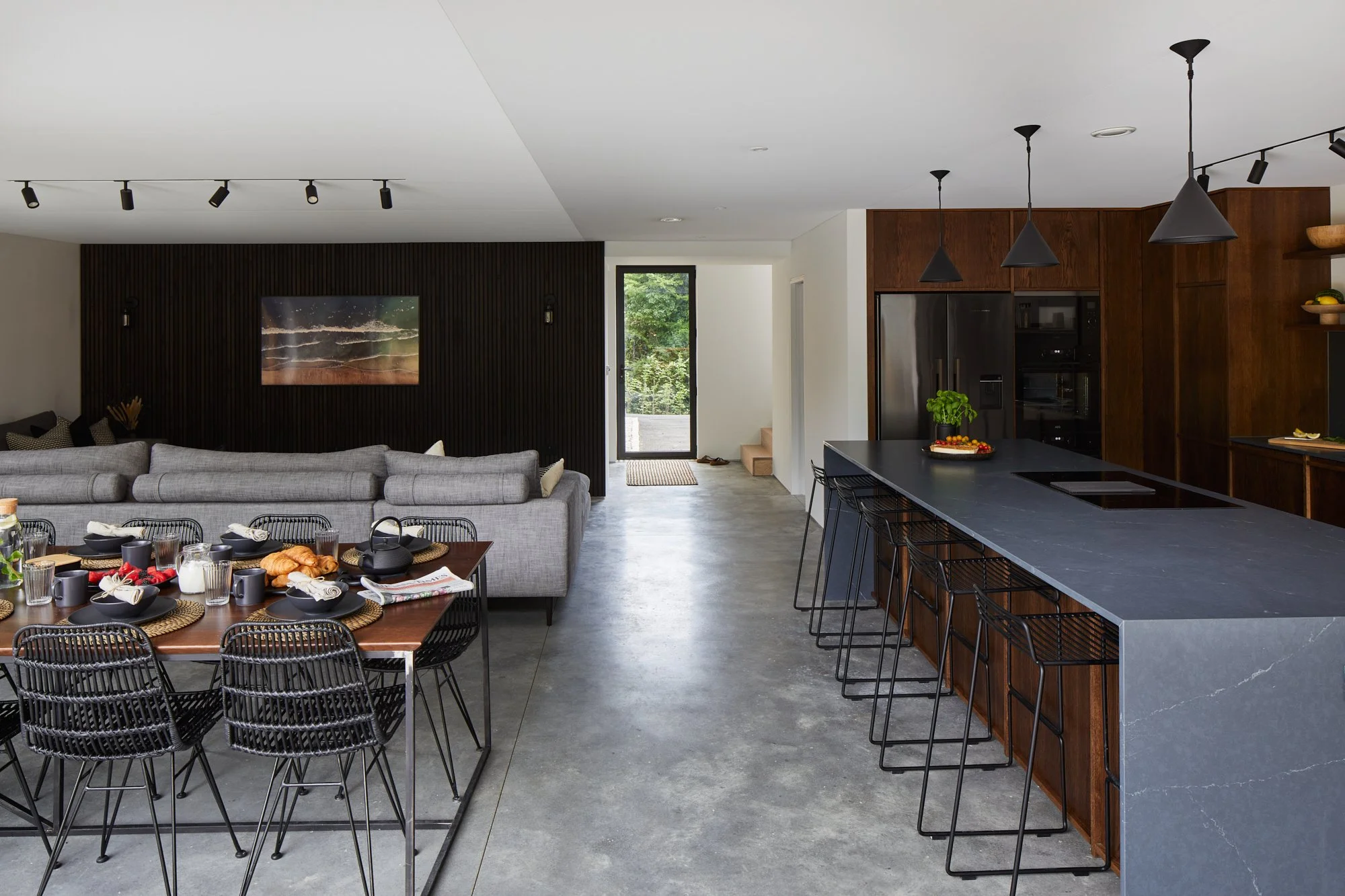 Open-concept kitchen and living room with dining table, gray sofa, black chairs, dark wood and black kitchen cabinetry, black island with barstools, pendant lighting, and sliding glass door showing greenery outside.
