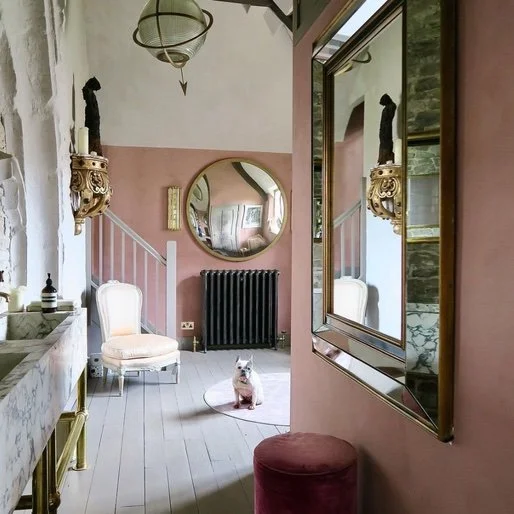 A stylish interior room with pink and white walls, a large mirror, a white upholstered chair, a small dog sitting on a round rug, and decorative elements including wall sconces, a round mirror, and wall art.