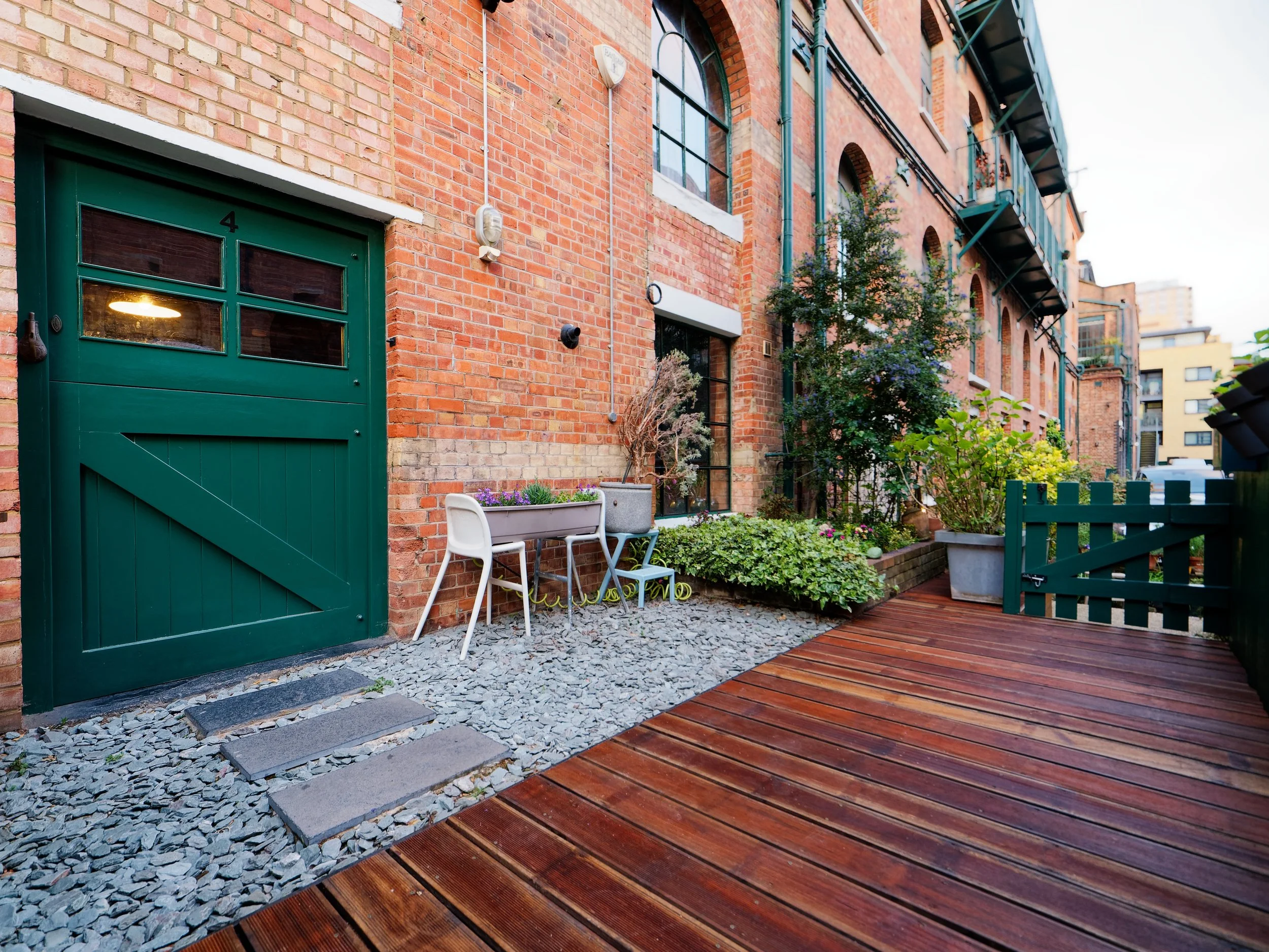 Outdoor patio area with a brick building, green door, potted plants, garden furniture, and wooden deck.