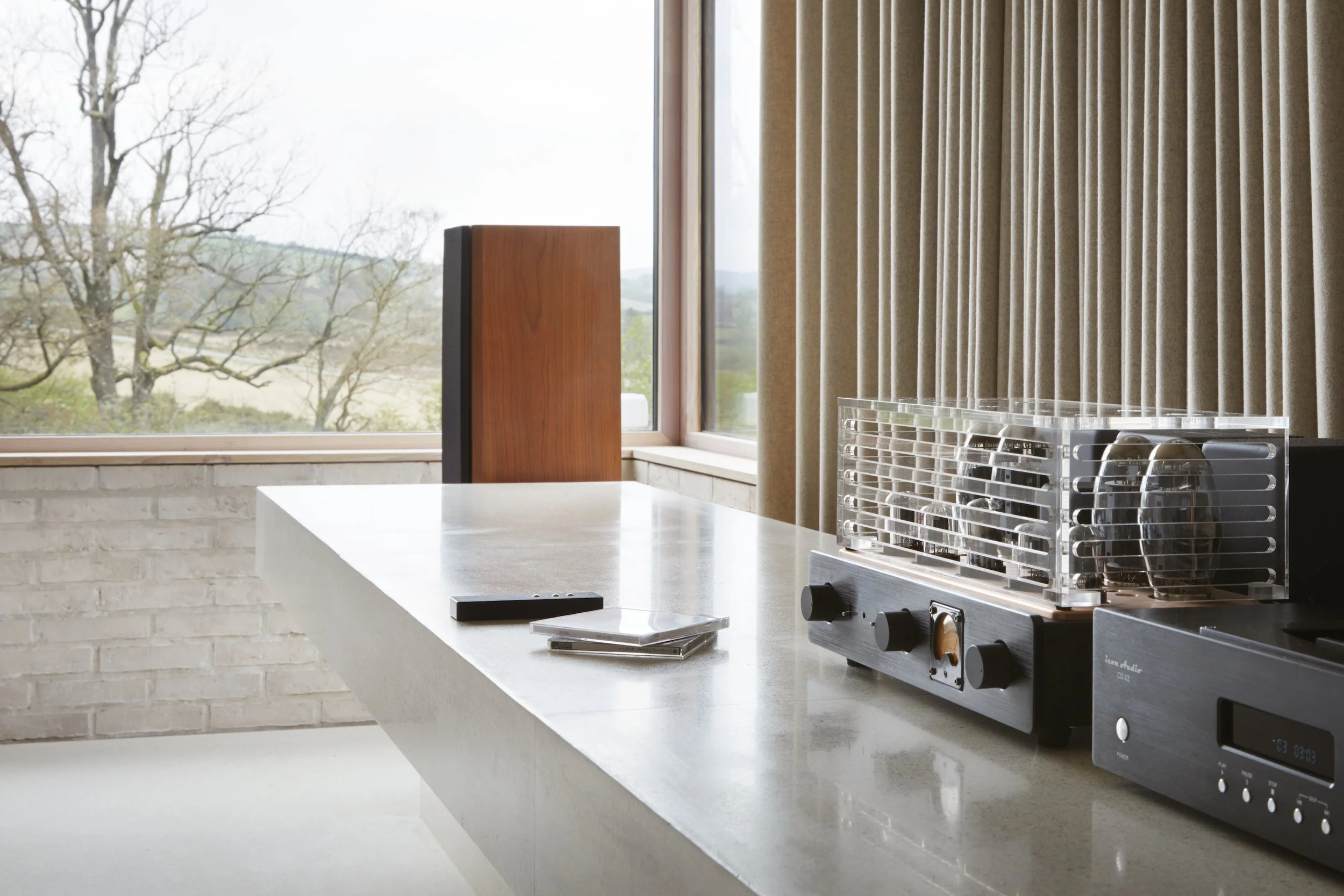 A modern kitchen countertop with a vintage radio, a remote, a notebook, and a CD case; a large window with beige curtains and a view of trees and sky outside.