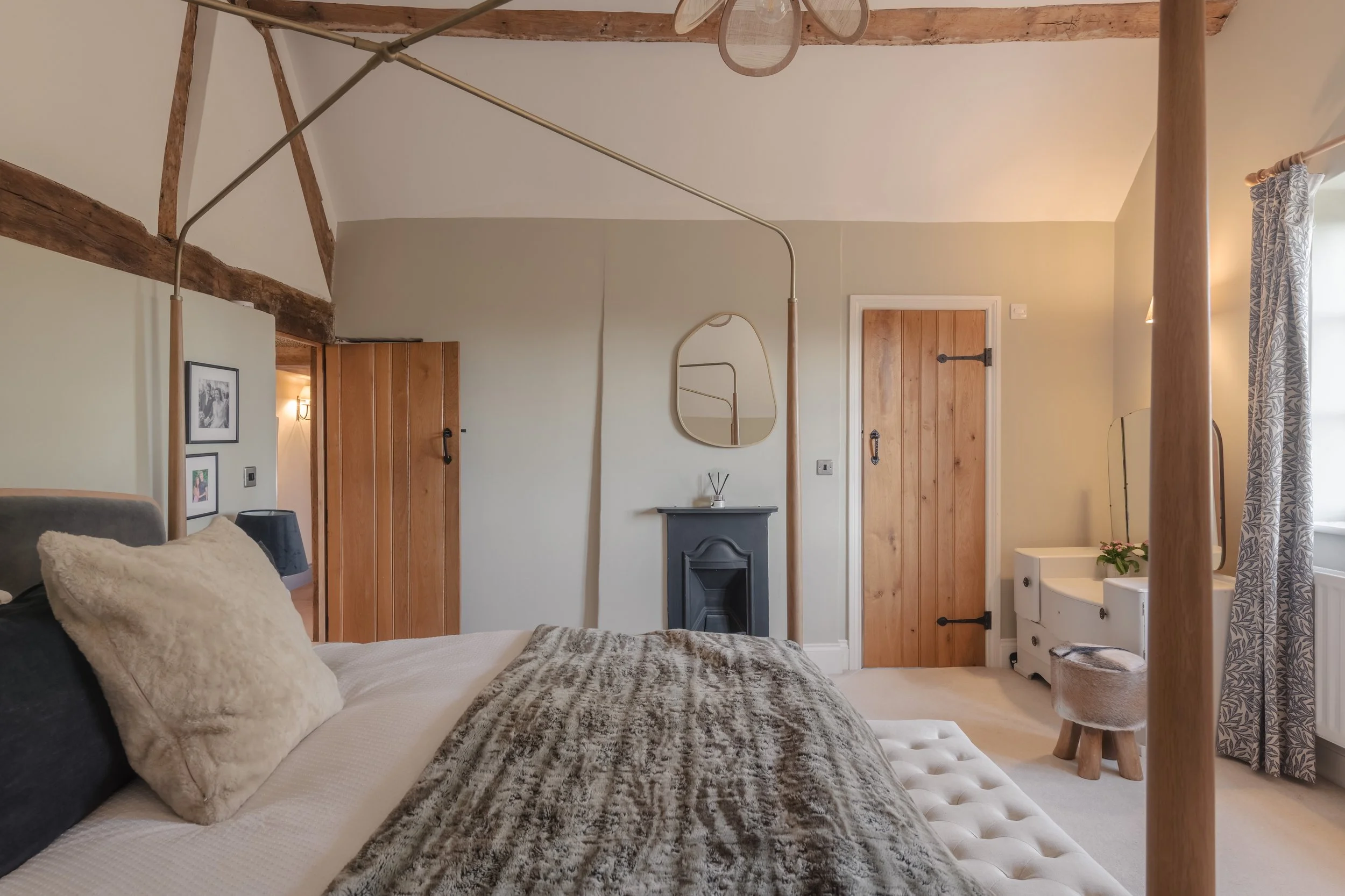 A cozy bedroom with a four-poster bed, plush bedding, and a textured pillow. The room features wooden doors, a small black fireplace with a round mirror above it, and a vanity with a mirror and flower pot. A window with patterned curtains allows natu