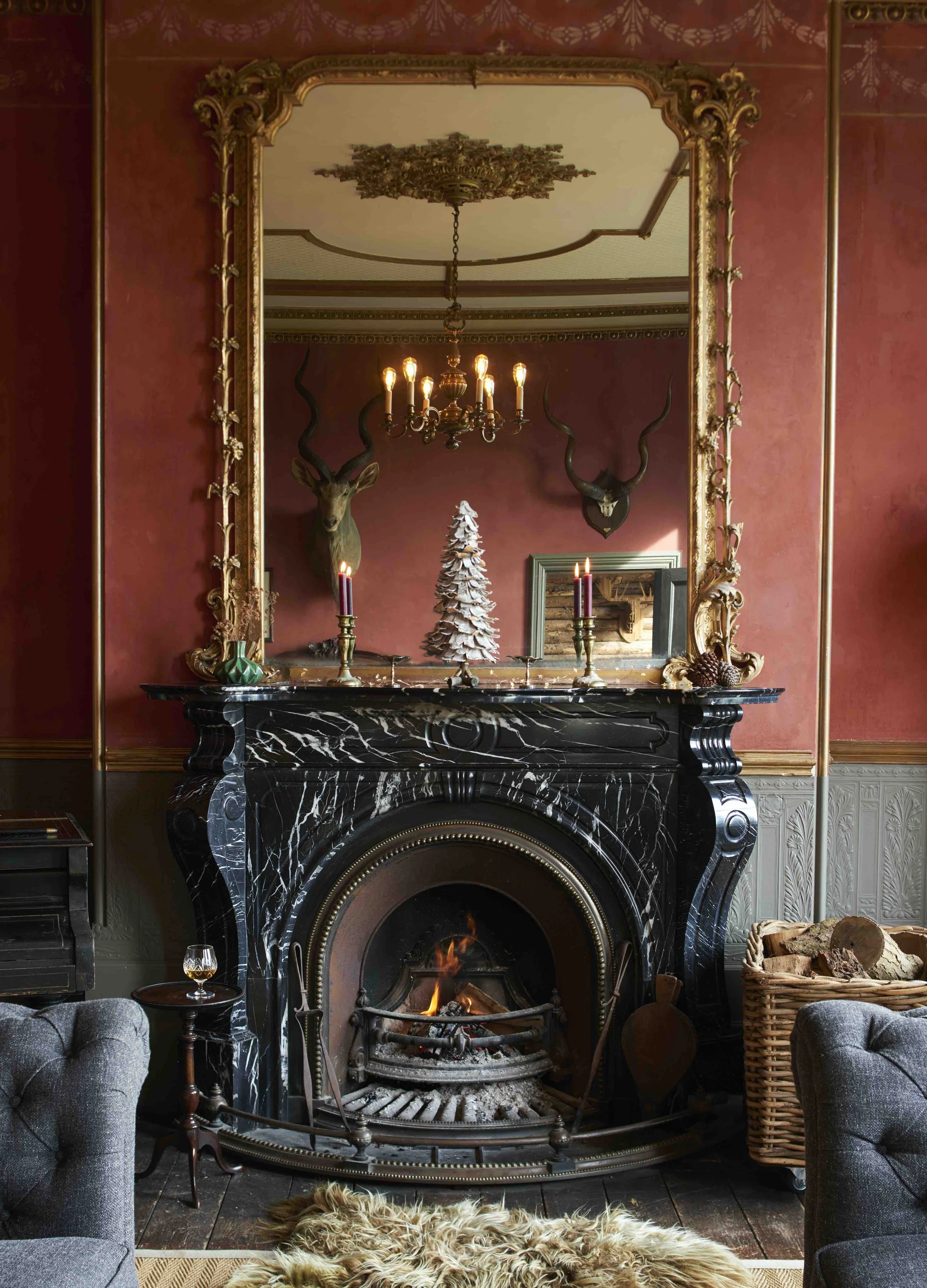 A vintage living room with a black marble fireplace, lit fire, and an ornate large mirror above it. The mantel holds a small Christmas tree, two purple candles, and decorative items. The red wall in the background features mounted antelope heads, a c