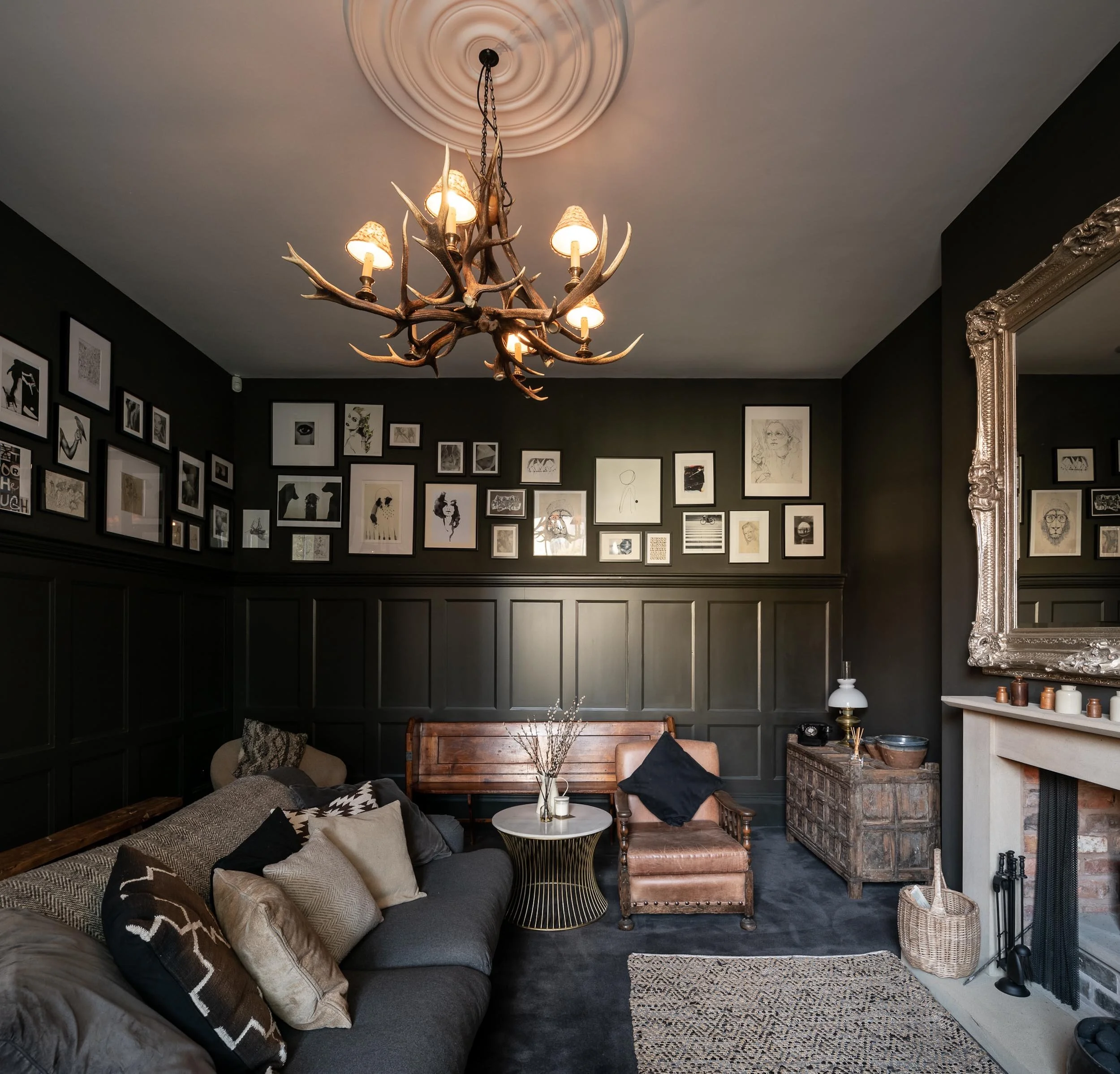Living room with black walls, gallery wall of framed artwork, dark furniture, chandelier with antlers, and a large decorative mirror.