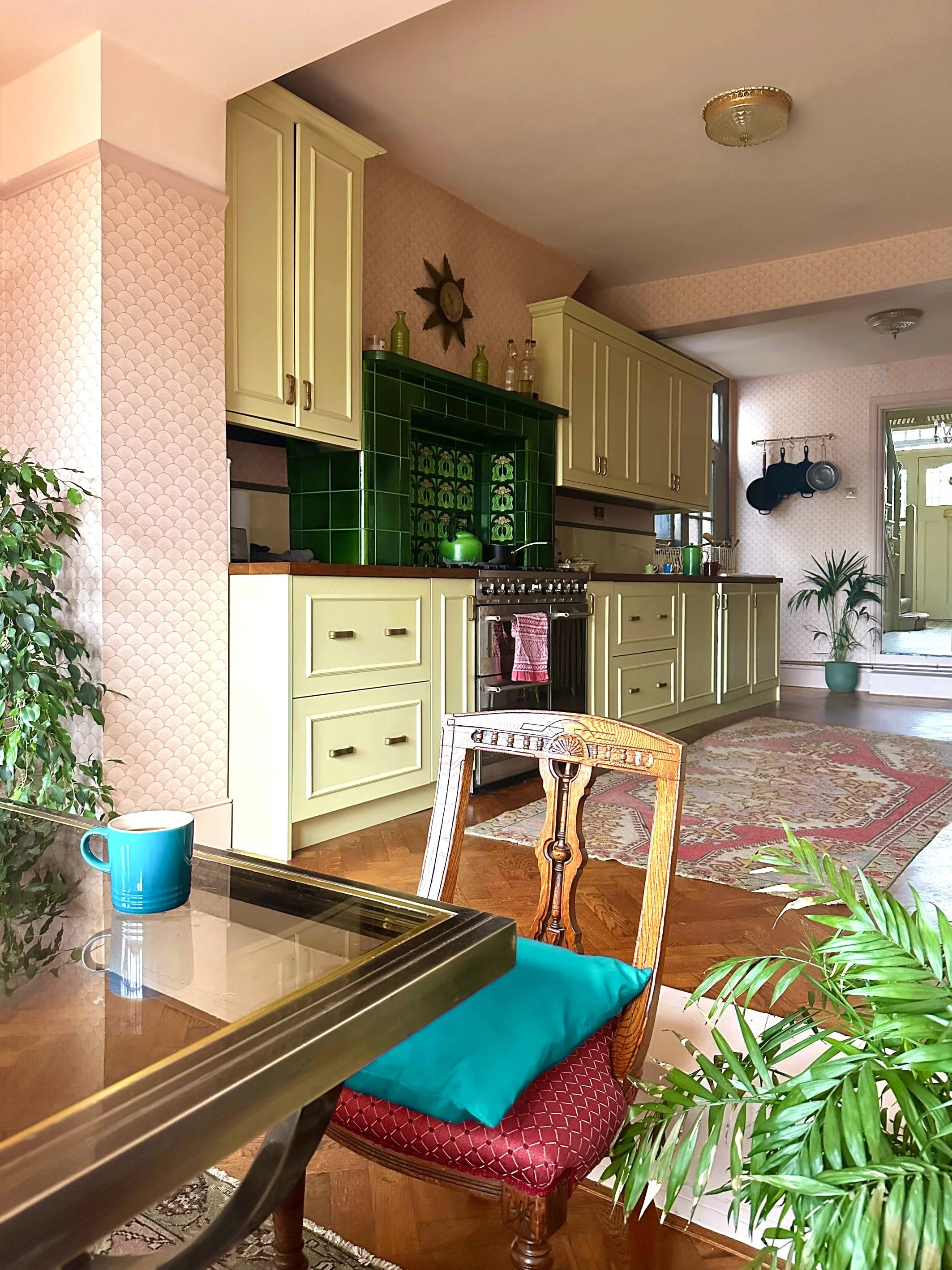 A cozy kitchen with green cabinets, a vintage stove, and a pink wallpaper. There is a dining table with a blue pillow on a wooden chair, a teal mug, and green plants. The area features wooden flooring and a patterned rug.