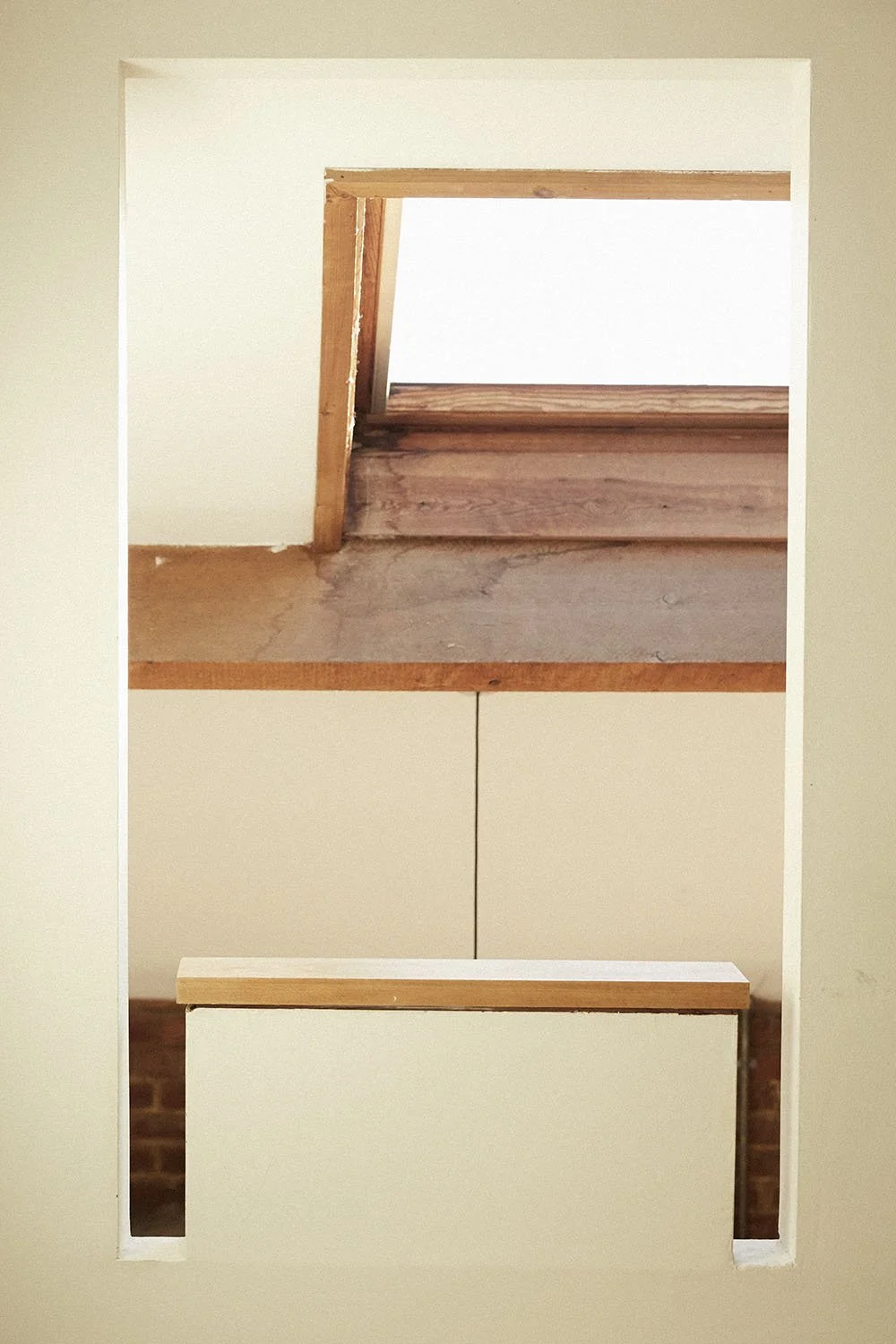 View through a rectangular opening in a wall showing a small wooden staircase and a skylight above.