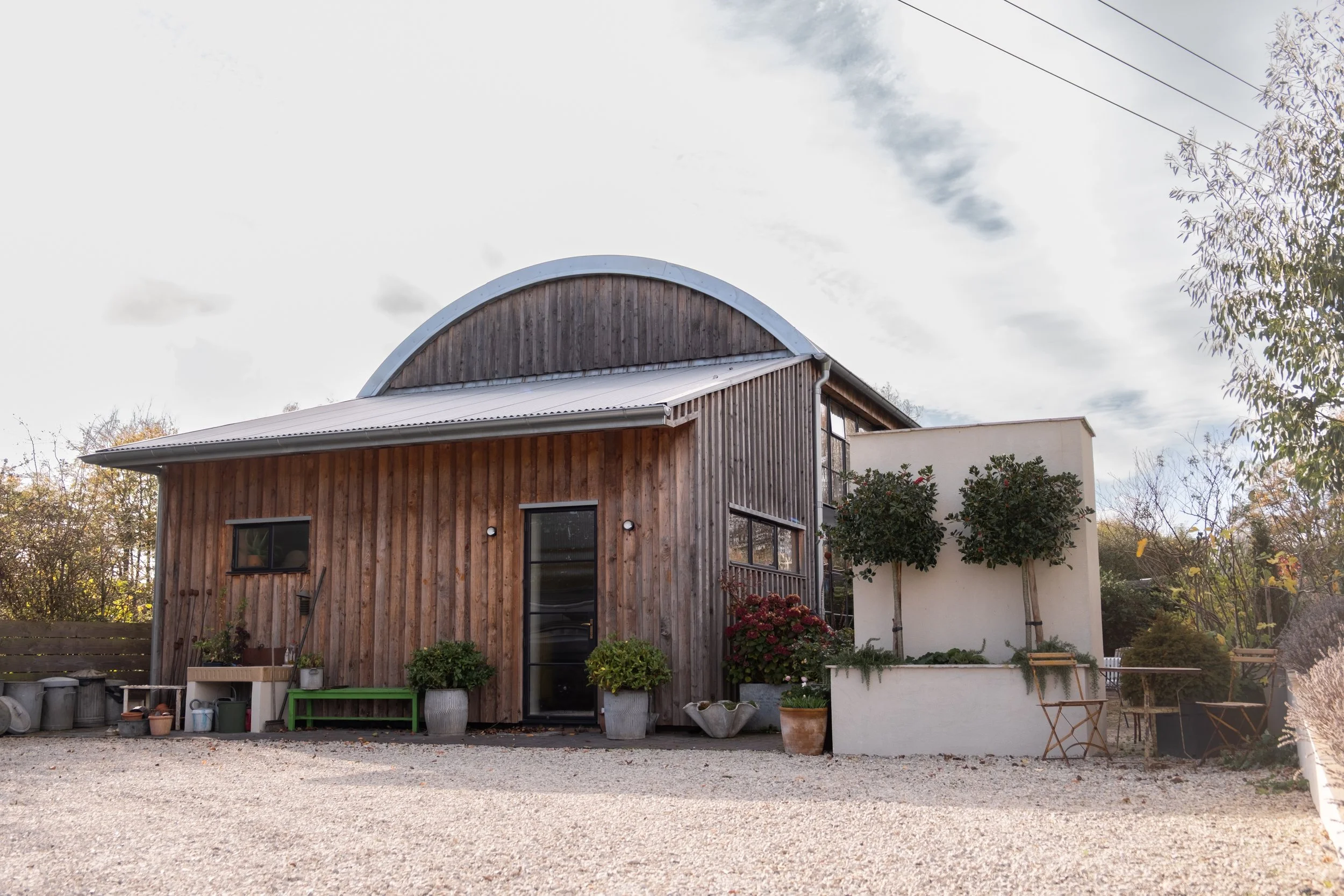 A modern house with wooden siding, large glass doors, and a rounded metal roof section, surrounded by plants and trees, with a gravel yard in the foreground and a cloudy sky above.