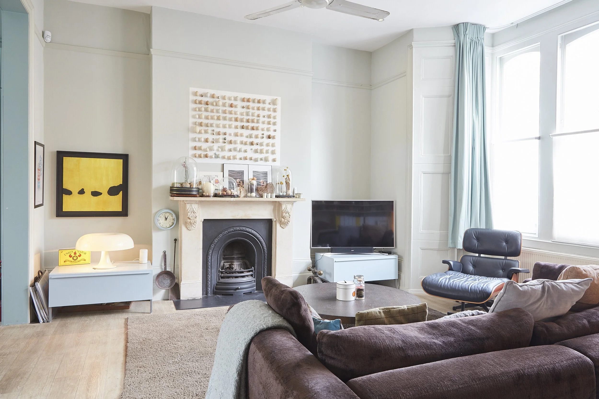 Interior of a living room with a brown sofa, a coffee table, a black lounge chair, and a TV on a stand. There is a fireplace with decorative items on the mantle and a colorful artwork on the wall. Large windows with light blue curtains allow natural 