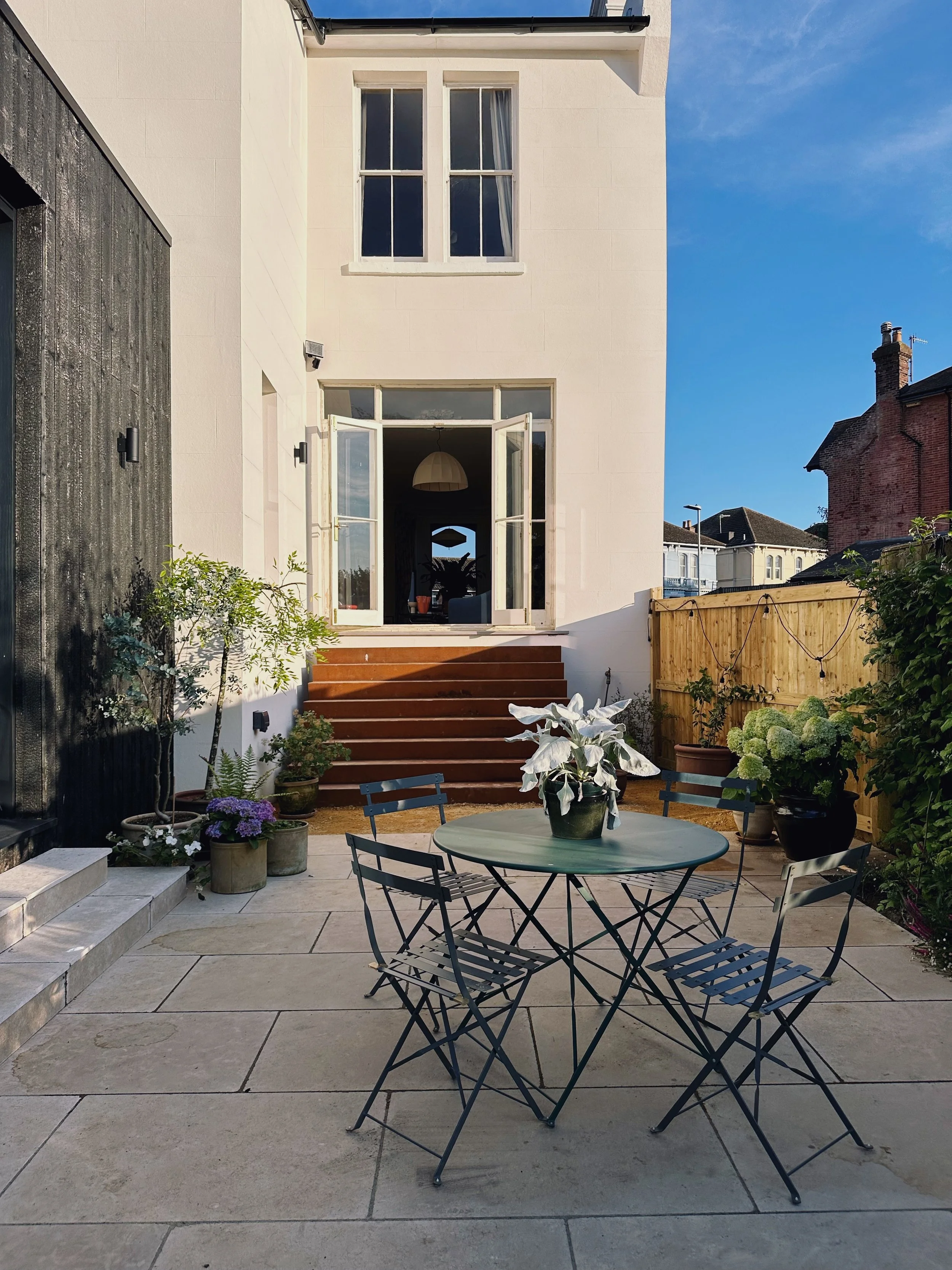 A backyard patio with a round metal table and four matching chairs, a potted plant with white leaves on the table, and various potted plants along the sides. The patio has wide stone tiles and steps leading up to a house with an open door, showing a 