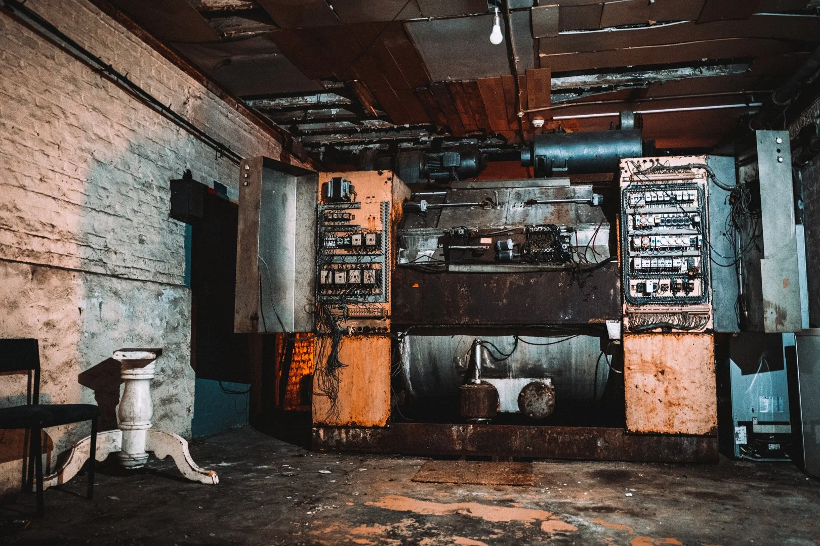 An abandoned room with exposed brick wall, old electrical panels with wires, and decaying ceiling panels with hanging light bulb.