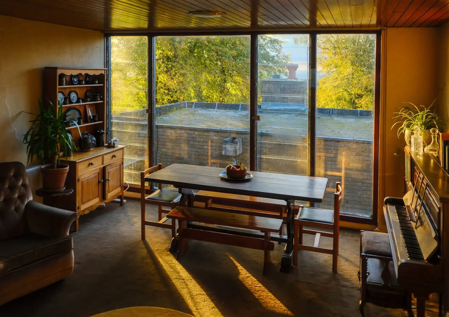 Living room with large window showing trees outside, a wooden dining table with four chairs, a piano on the right, a cabinet with plates on the left, and indoor plants, bathed in warm sunlight.