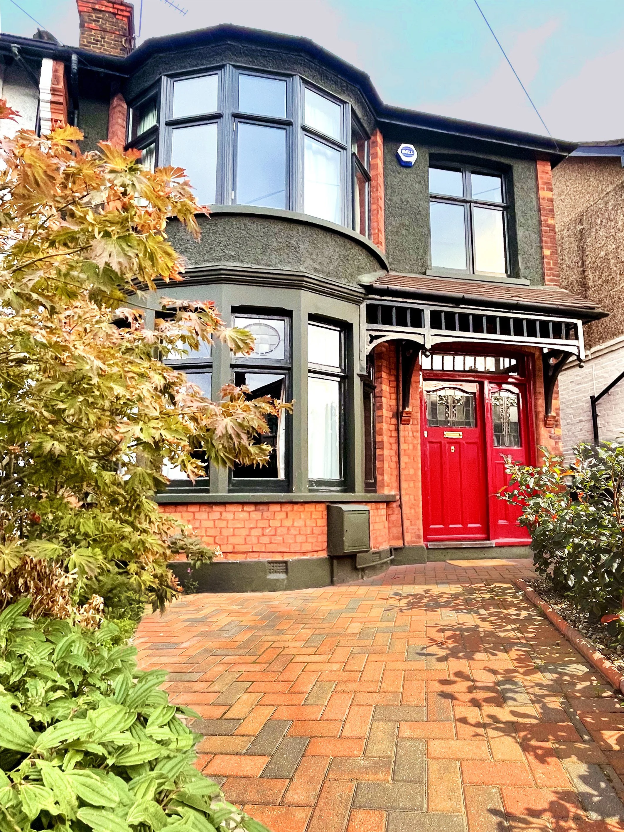 Red front door of a brick house with green framing and bay windows, surrounded by plants and a brick walkway.