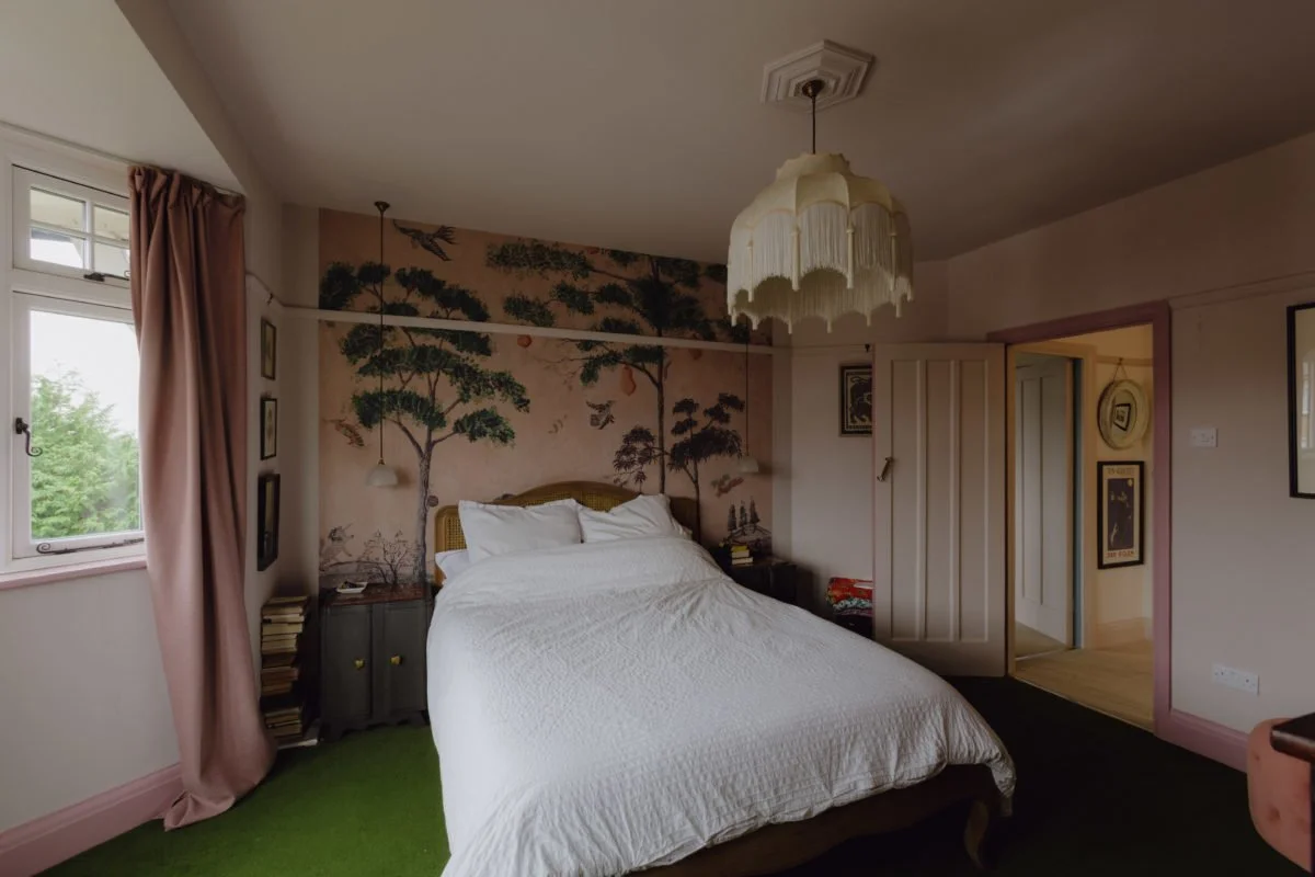 A bedroom with a white bed, pink curtains, and a mural of trees and birds on the wall behind the bed. There are nightstands on either side of the bed with books and small lamps. A vintage-style chandelier hangs from the ceiling. The room has a small 