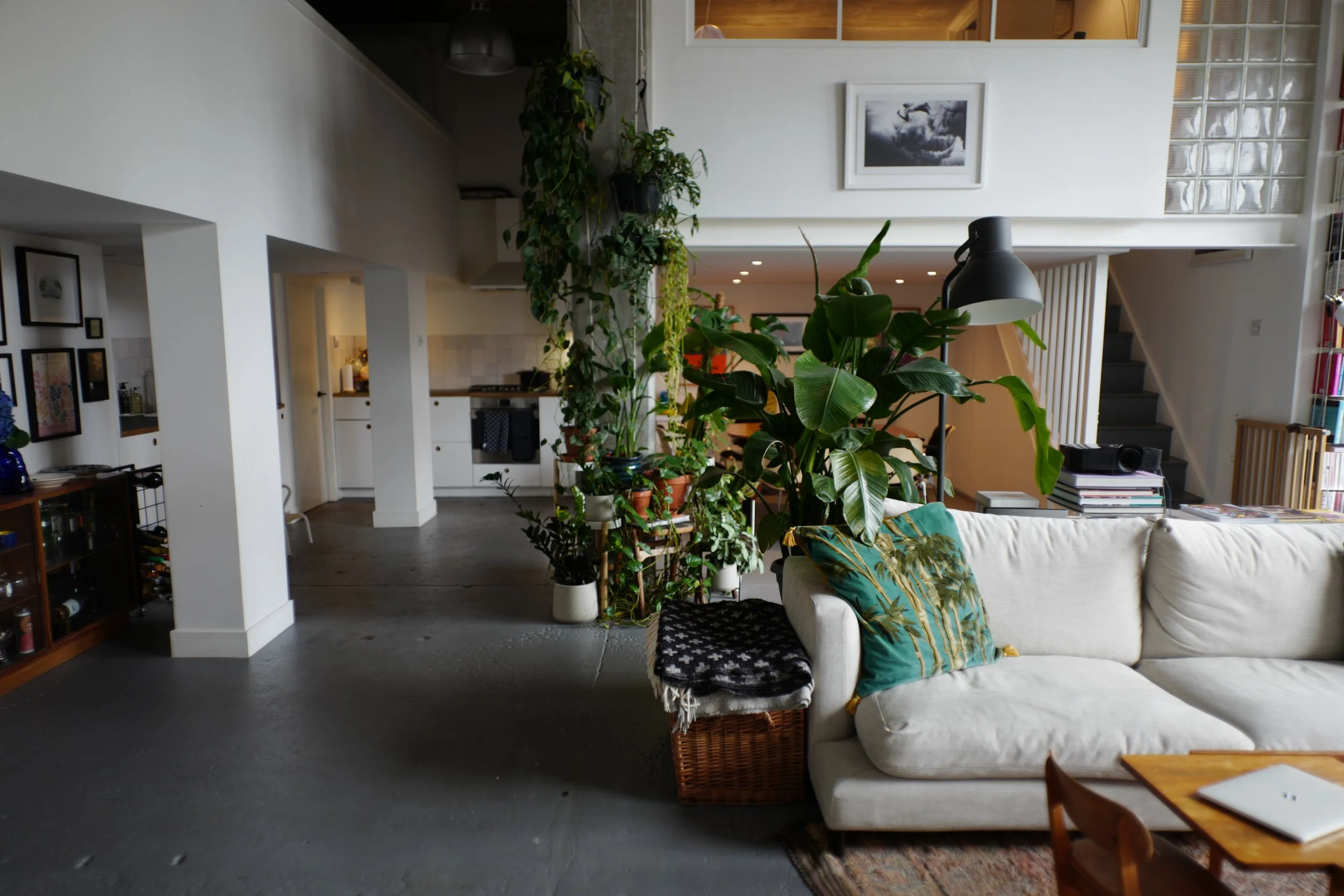 Living room with a white sofa, green plants, a wooden coffee table, cushions with tropical print, a black lamp, and a bookshelf. Kitchen area visible in the background.