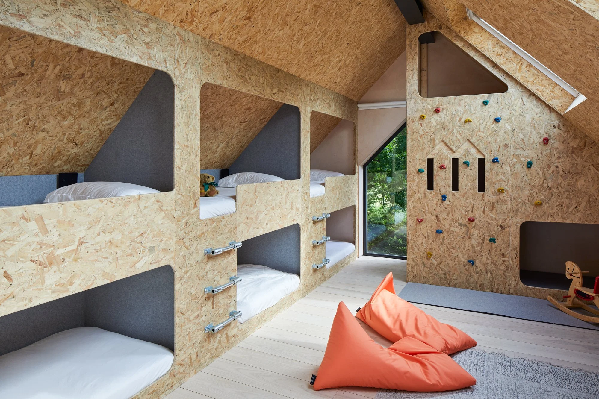 Interior of a children's bedroom with wooden bunk beds, a climbing wall, a rocking horse, and bean bag chairs, all under a sloped ceiling with large skylight window.