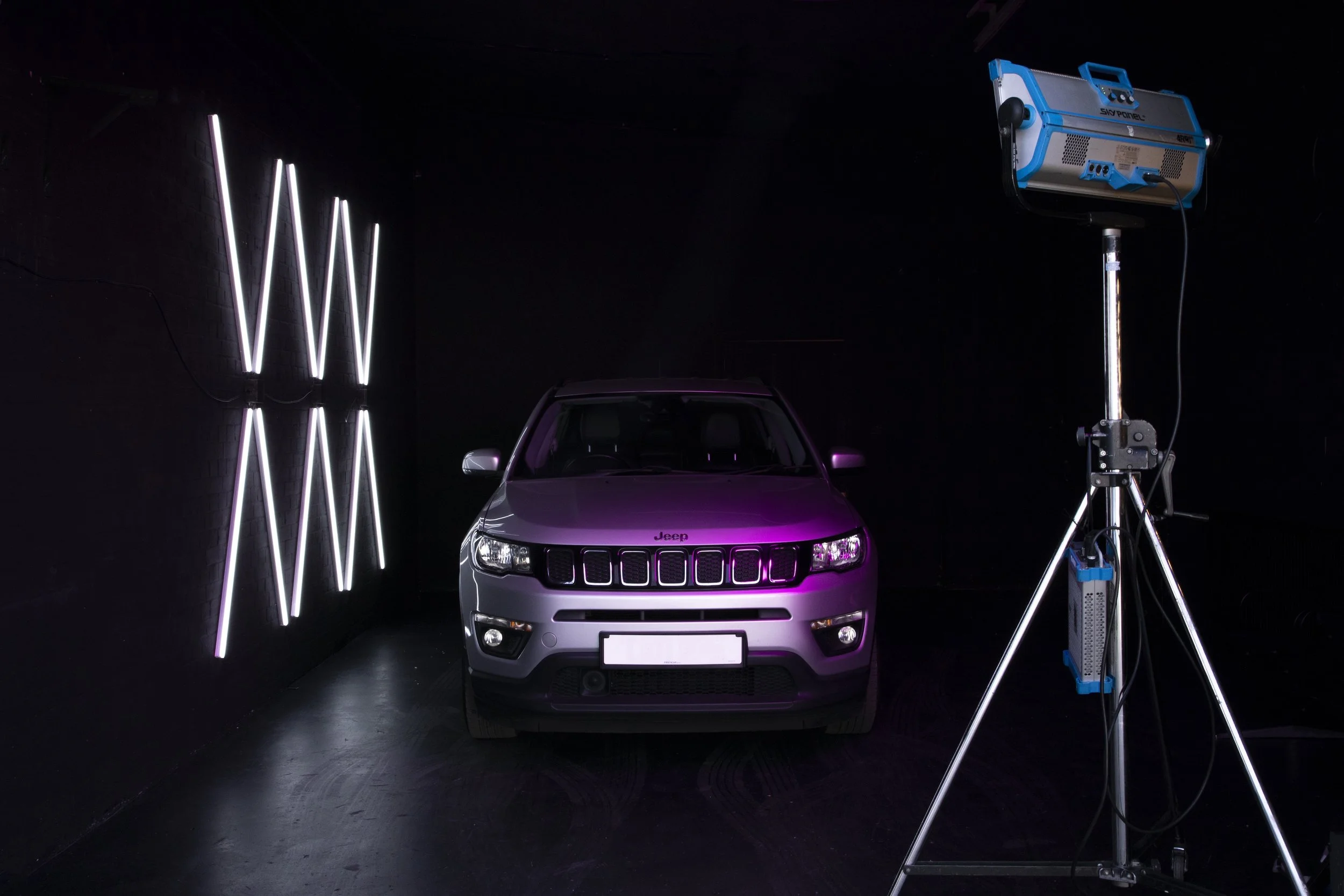 A silver Jeep SUV is positioned in a dark room with a black background, illuminated by a purple light. To the right, there is a large blue and silver lighting equipment on a stand, and on the left, vertical neon lights on the wall.