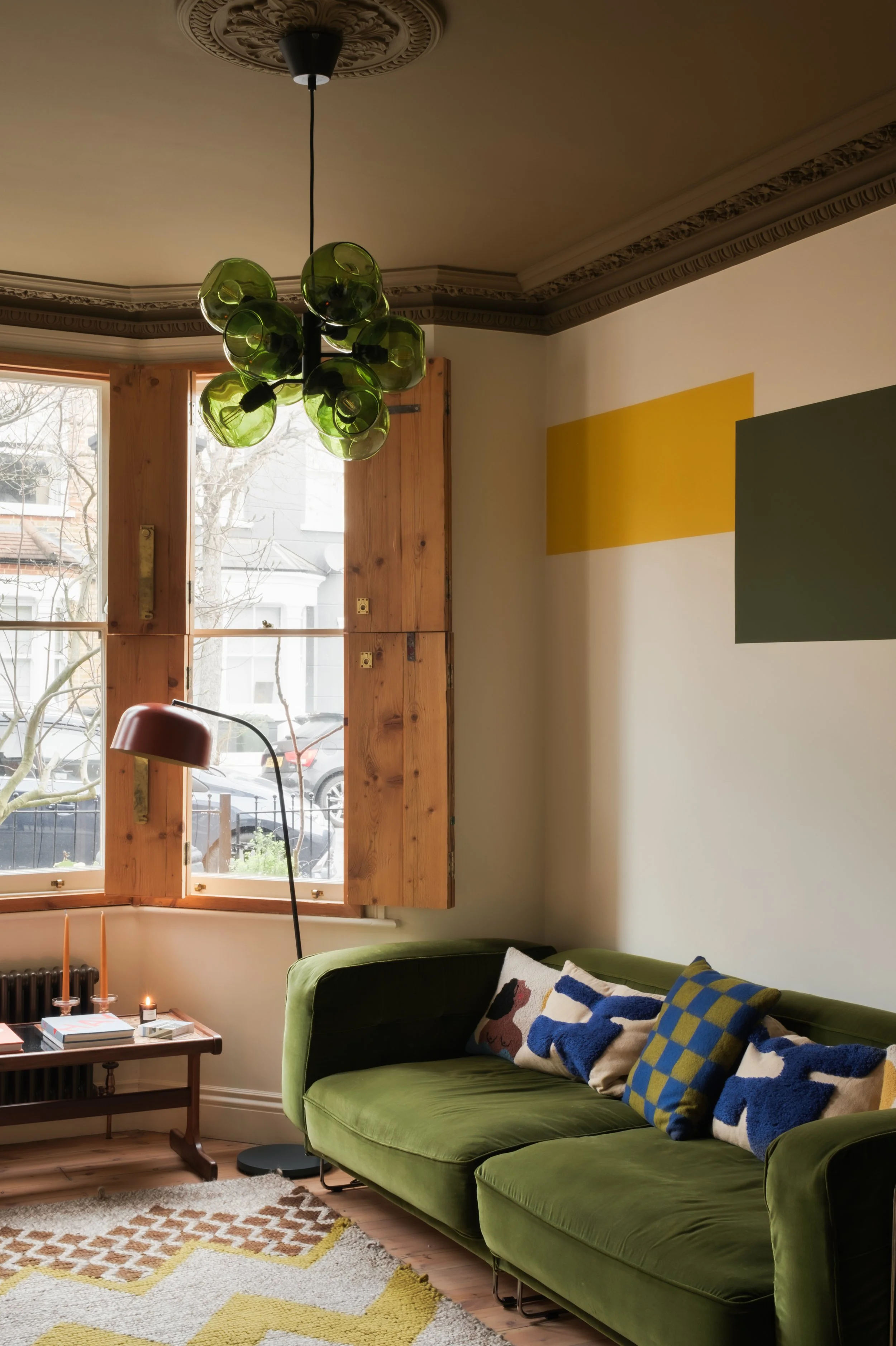 Living room with green velvet sofa, colorful patterned cushions, wooden window shutters, a window showing trees and parked cars outside, a patterned rug, a floor lamp, and a modern ceiling light fixture.
