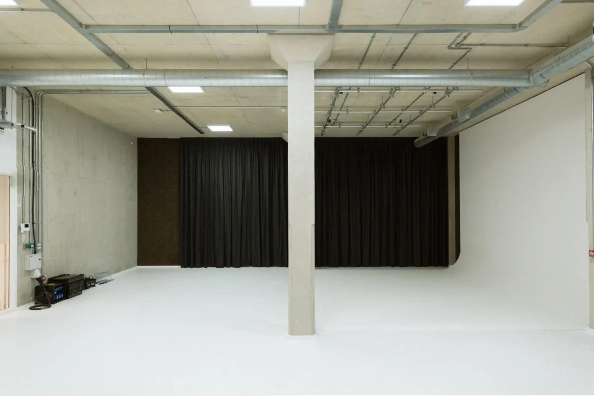 Empty indoor space with a stage curtain, concrete walls, and ceiling with exposed pipes and ducts.