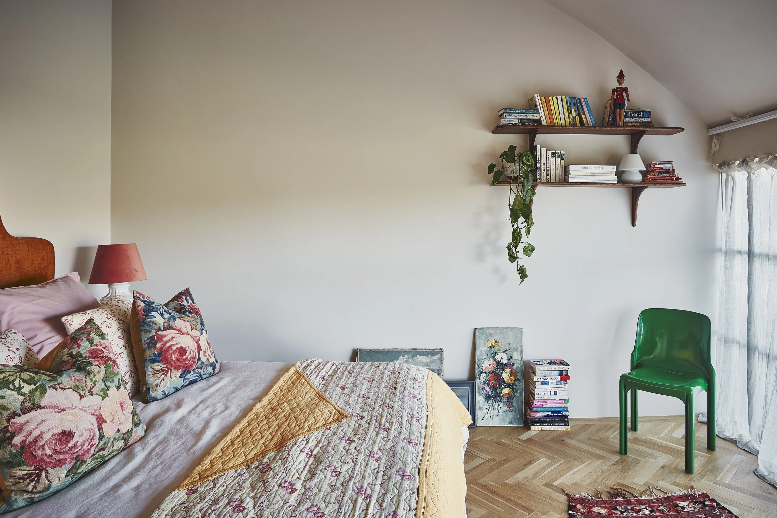 A cozy bedroom corner with a bed decorated with floral and pink patterned pillows, a pink lampshade on a white bedside table, a green chair, and a wooden wall shelf filled with books, a white lamp, and a small wooden figurine, with natural light comi
