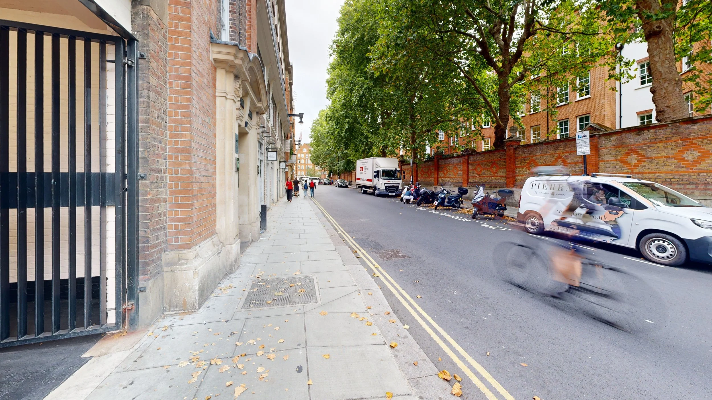 A city street with pedestrians walking on the sidewalk, a white delivery van and a moving truck on the road, and parked scooters against a brick wall. A cyclist is blurred in motion in the foreground.