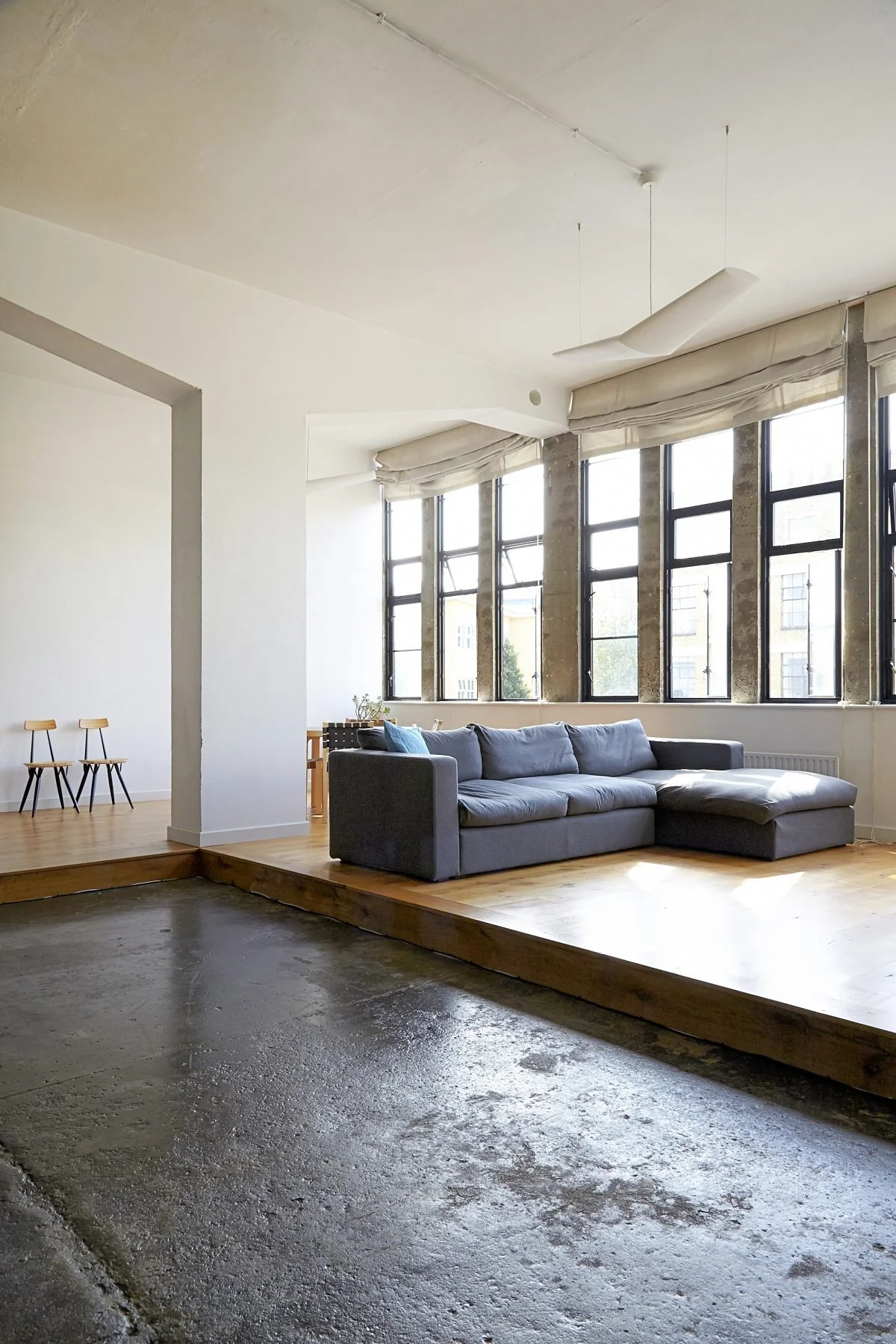 Living room with large windows, a gray sectional sofa, and a platform with wood flooring. The area has a partially unfinished look with exposed concrete and window shades draped over the windows.