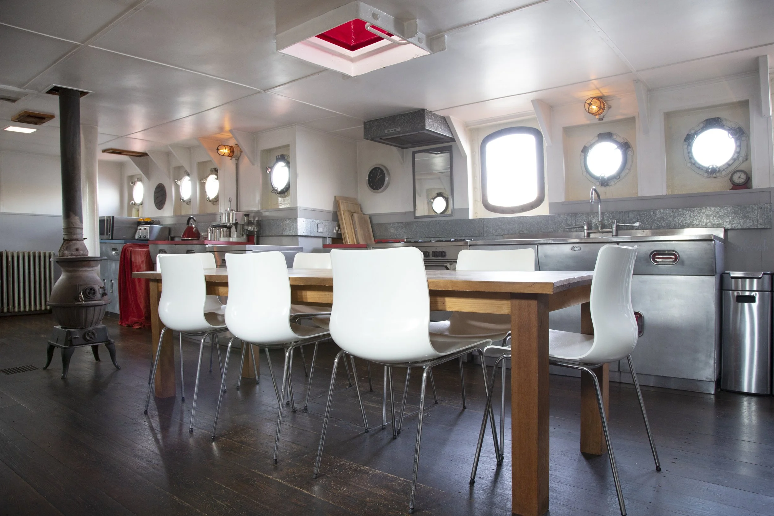 Empty dining area with white chairs around a wooden table, stainless steel kitchen counters, porthole windows, and a wood stove in a nautical-themed room.