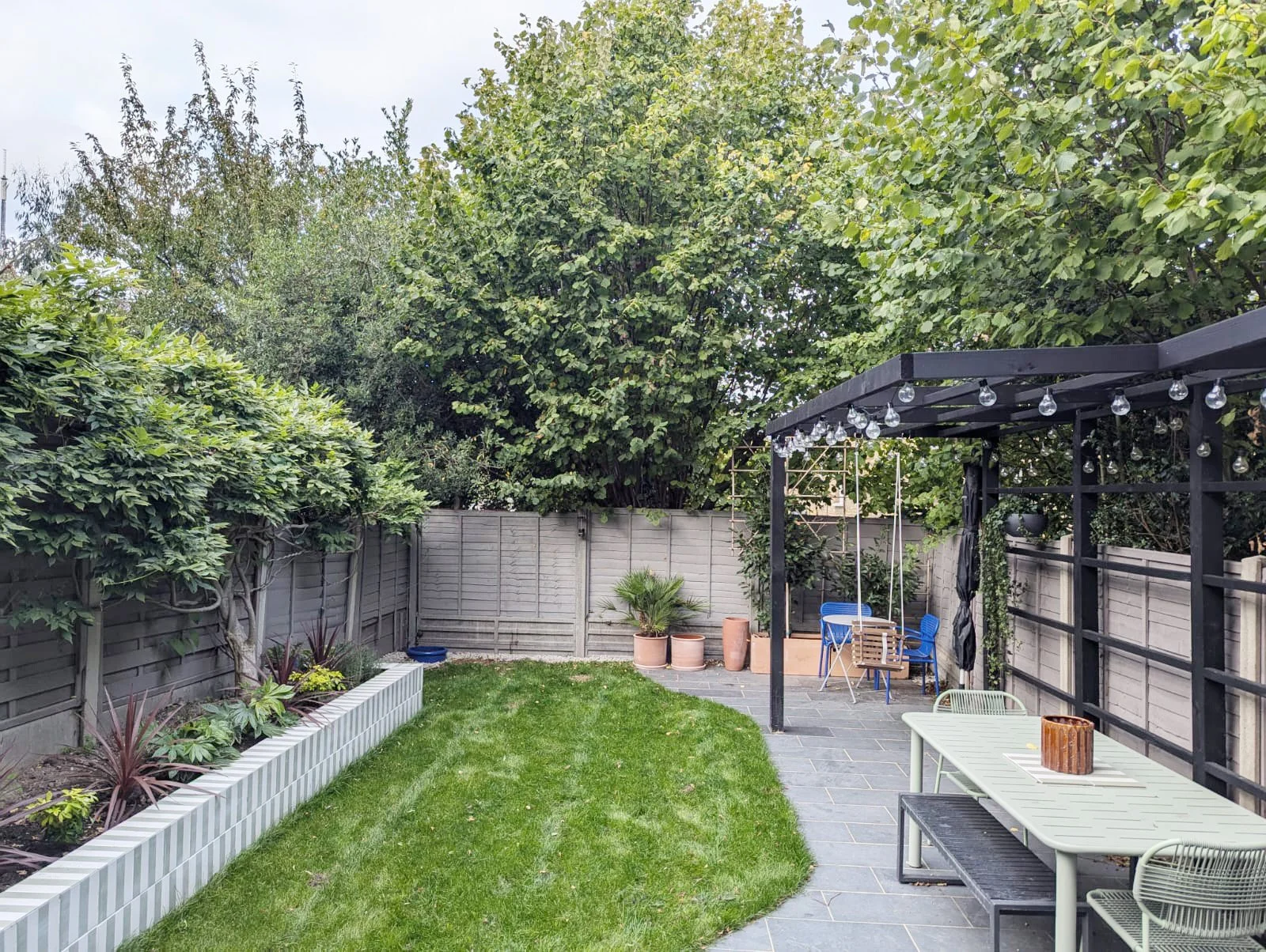 A backyard garden with a grassy lawn, potted plants, a gray wooden fence, and a black pergola with hanging string lights. There is outdoor furniture, including a table, chairs, and a swing.