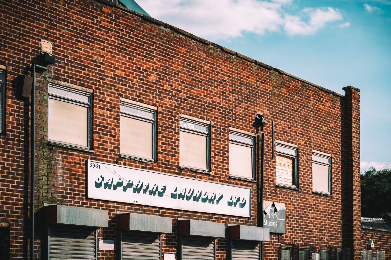 A brick building with four windows covered with blinds and shutters, a sign reading 'HAPPAGE LAUNDRY' and a small awning at the bottom.