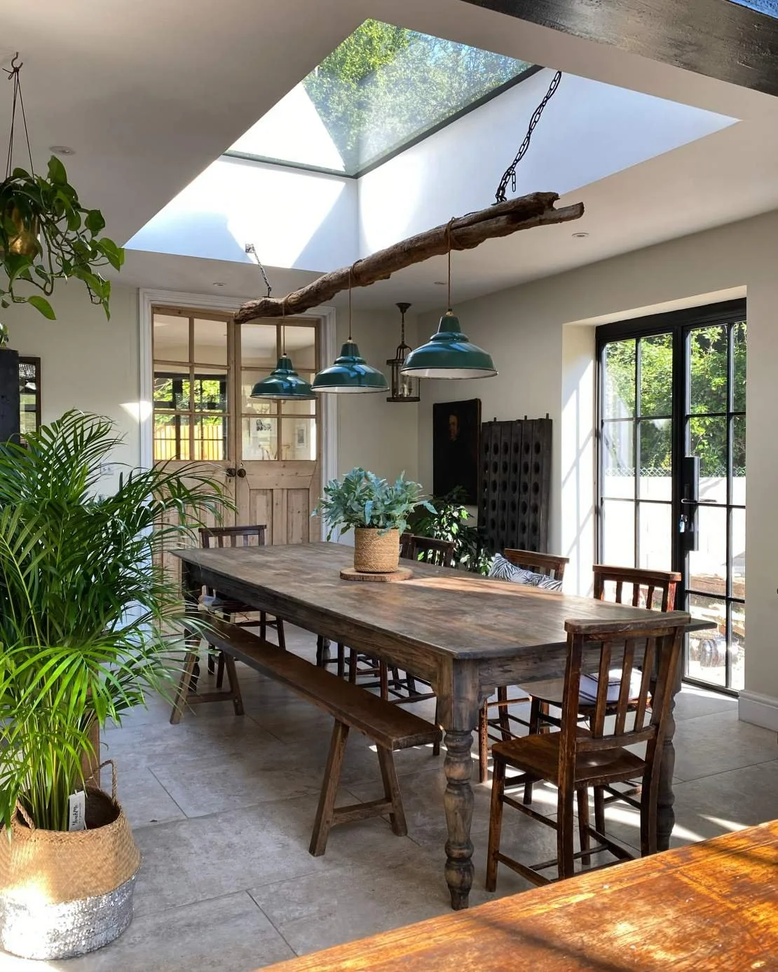 Bright dining area with a rustic wooden table, chairs, and a bench, illuminated by natural light from a large skylight and glass doors, decorated with plants and vintage-style pendant lights.