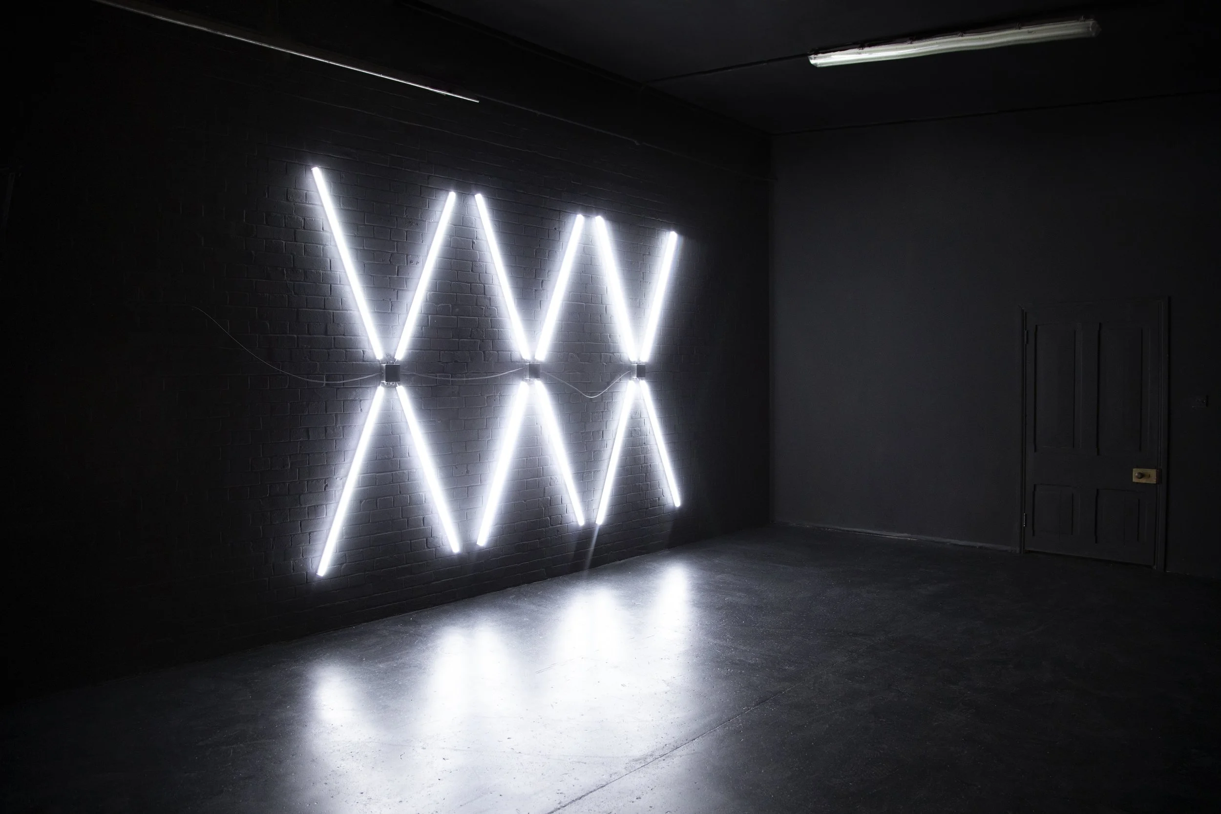 An interior room with black walls and ceiling, illuminated by modern white neon lights arranged in a repeating 'W' pattern on one wall. The floor is dark and reflective, with a closed door on the right side of the room.