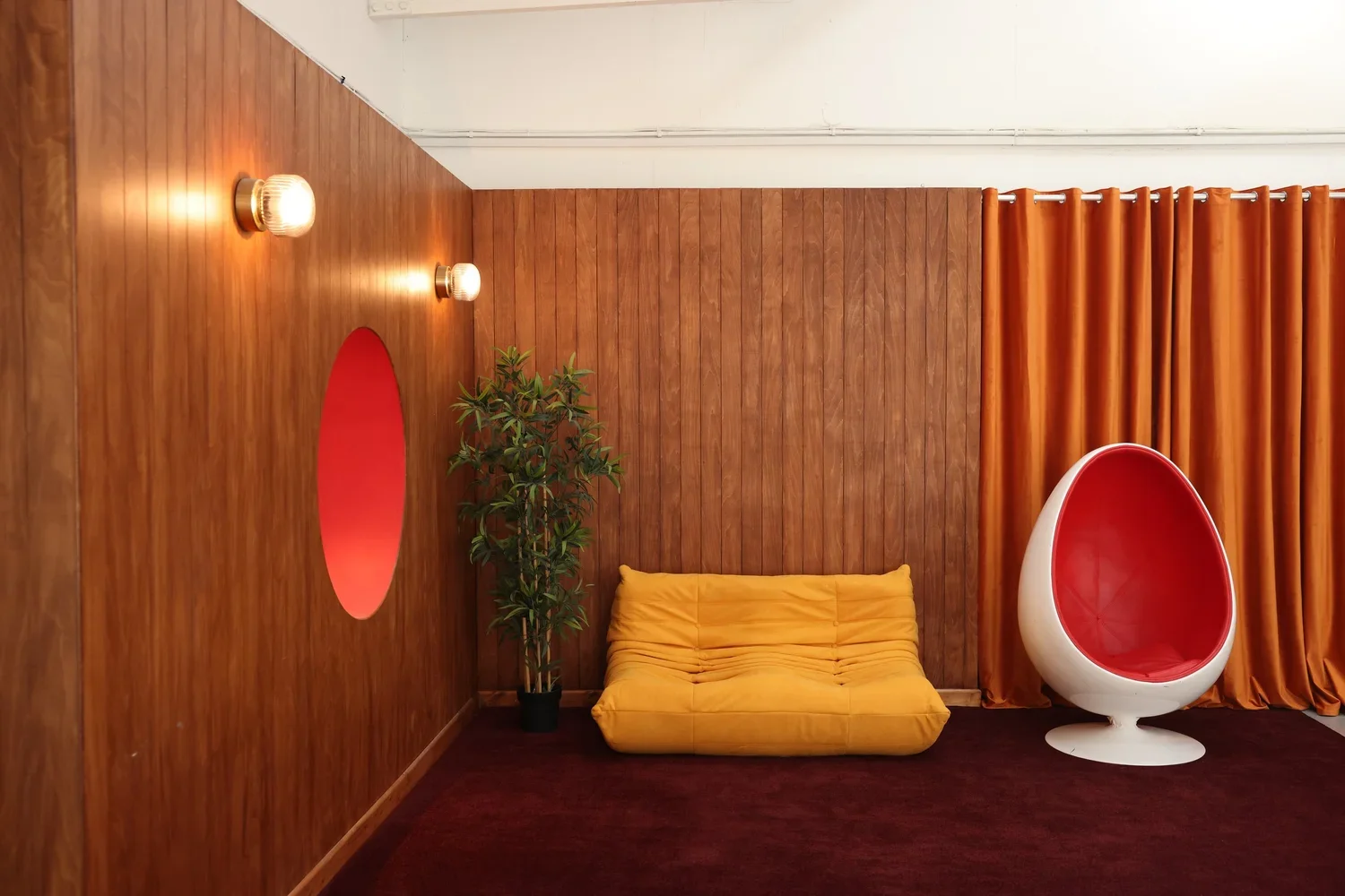 Retro photography studio with wood panelled walls, yellow cushioned seating, an orange curtain, a potted plant, and a white egg-shaped chair with red interior.