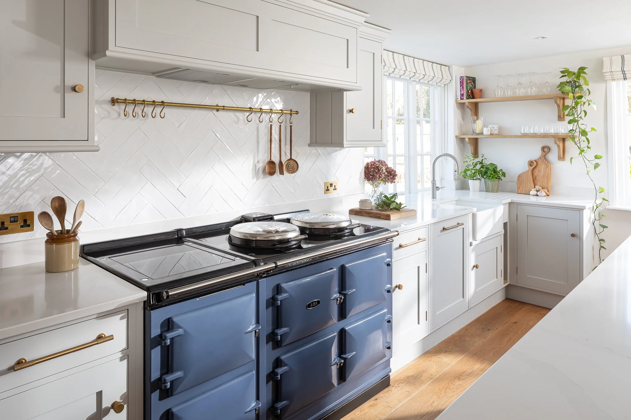 Bright kitchen with white cabinets, a blue vintage stove, open wooden shelves with glassware, plants, and kitchen utensils, and a window with striped curtains.
