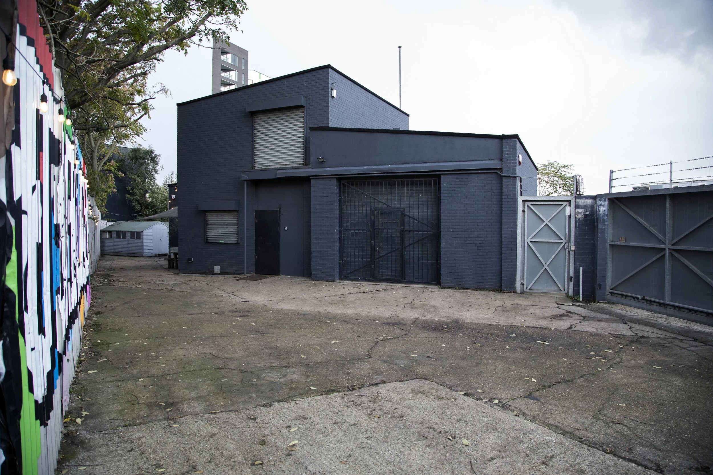 Side alley view of a modern black brick building with a gated entrance and a small fenced door, surrounded by a paved cracked driveway and a colorful graffiti-covered wall on the left, with trees and a partly cloudy sky in the background.