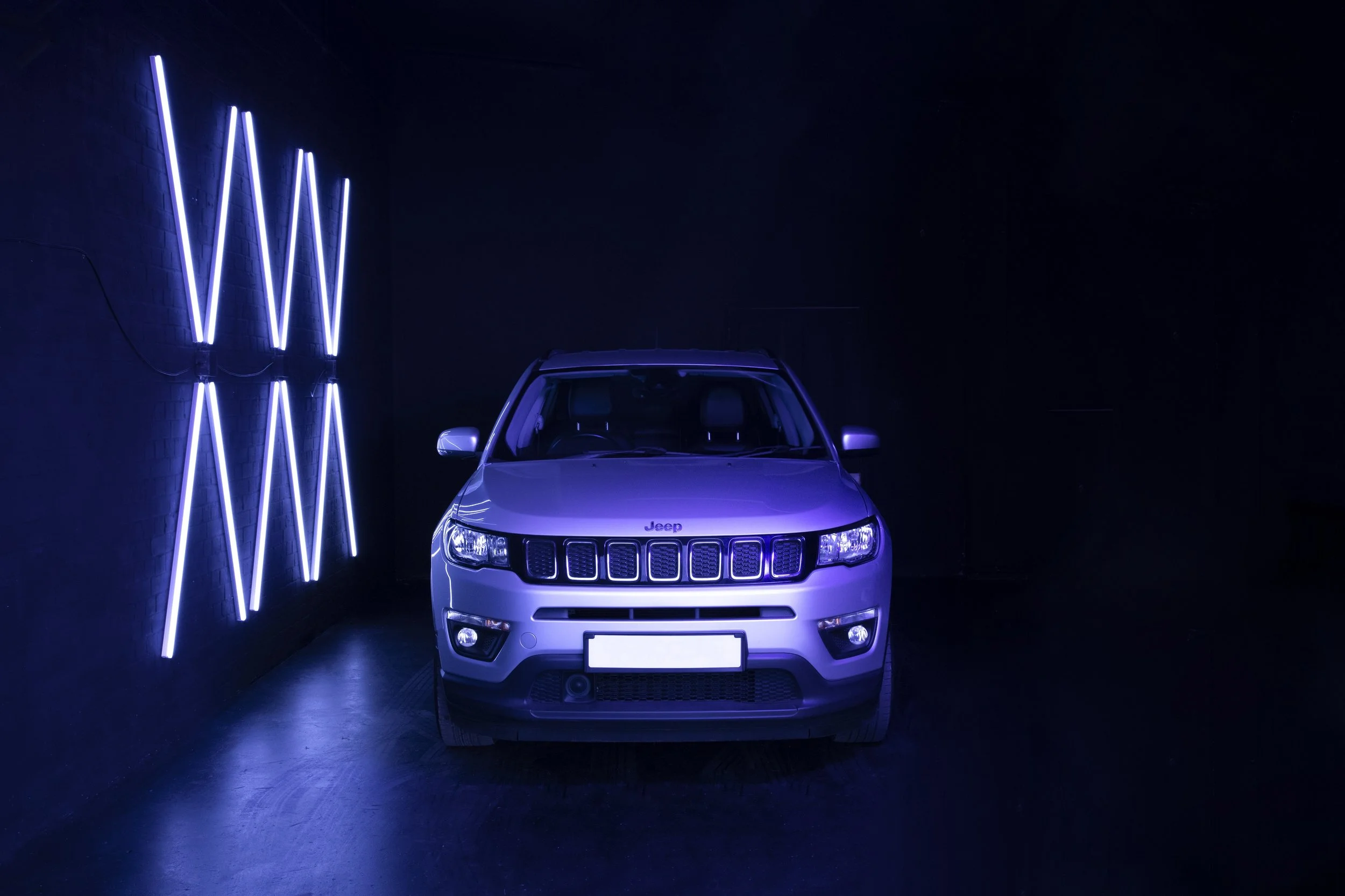Silver Jeep SUV parked in a dark room with a black wall, illuminated by blue neon lights on the left side.
