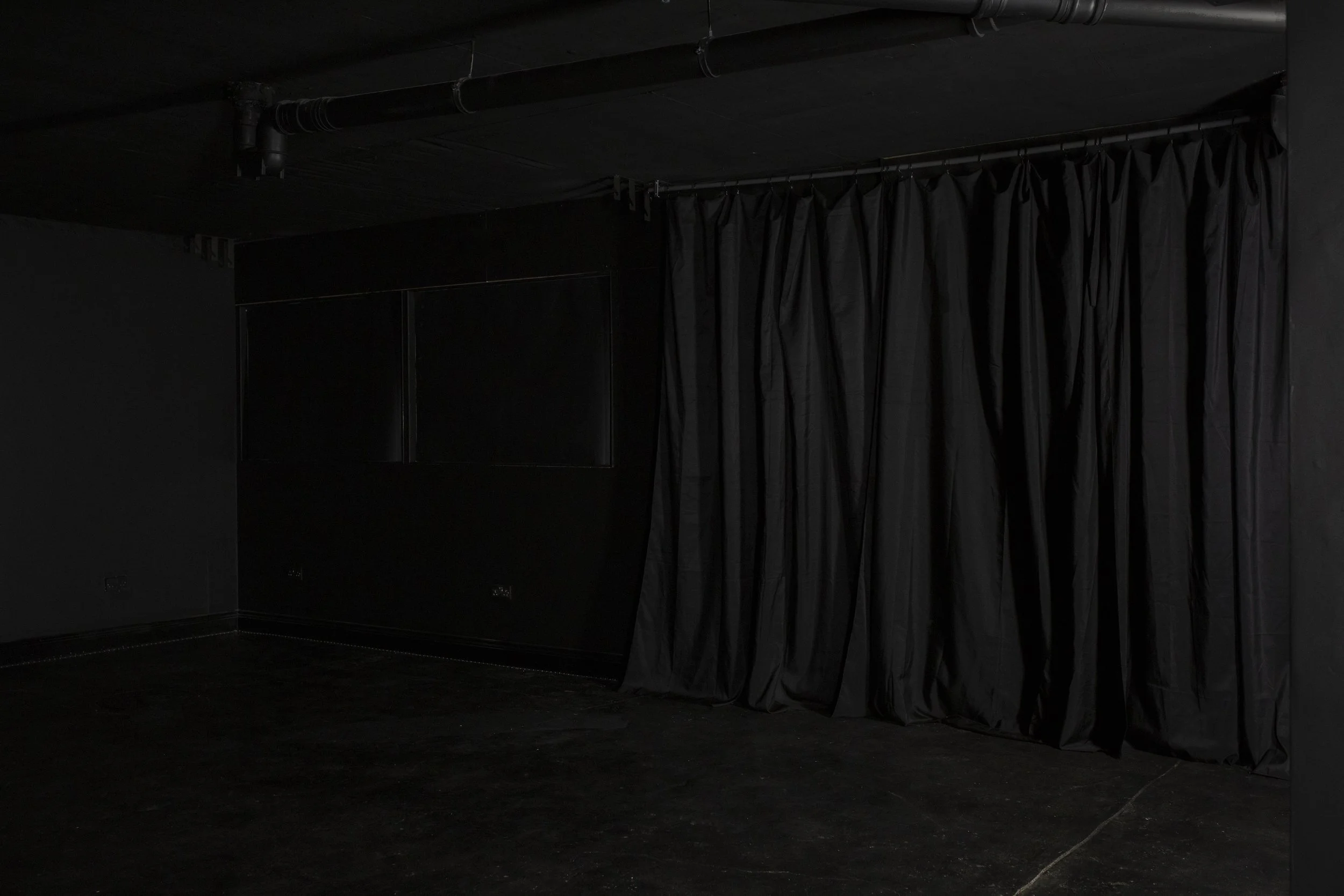 A dark empty stage or black box theater with closed black curtains on the right and black walls and ceiling.