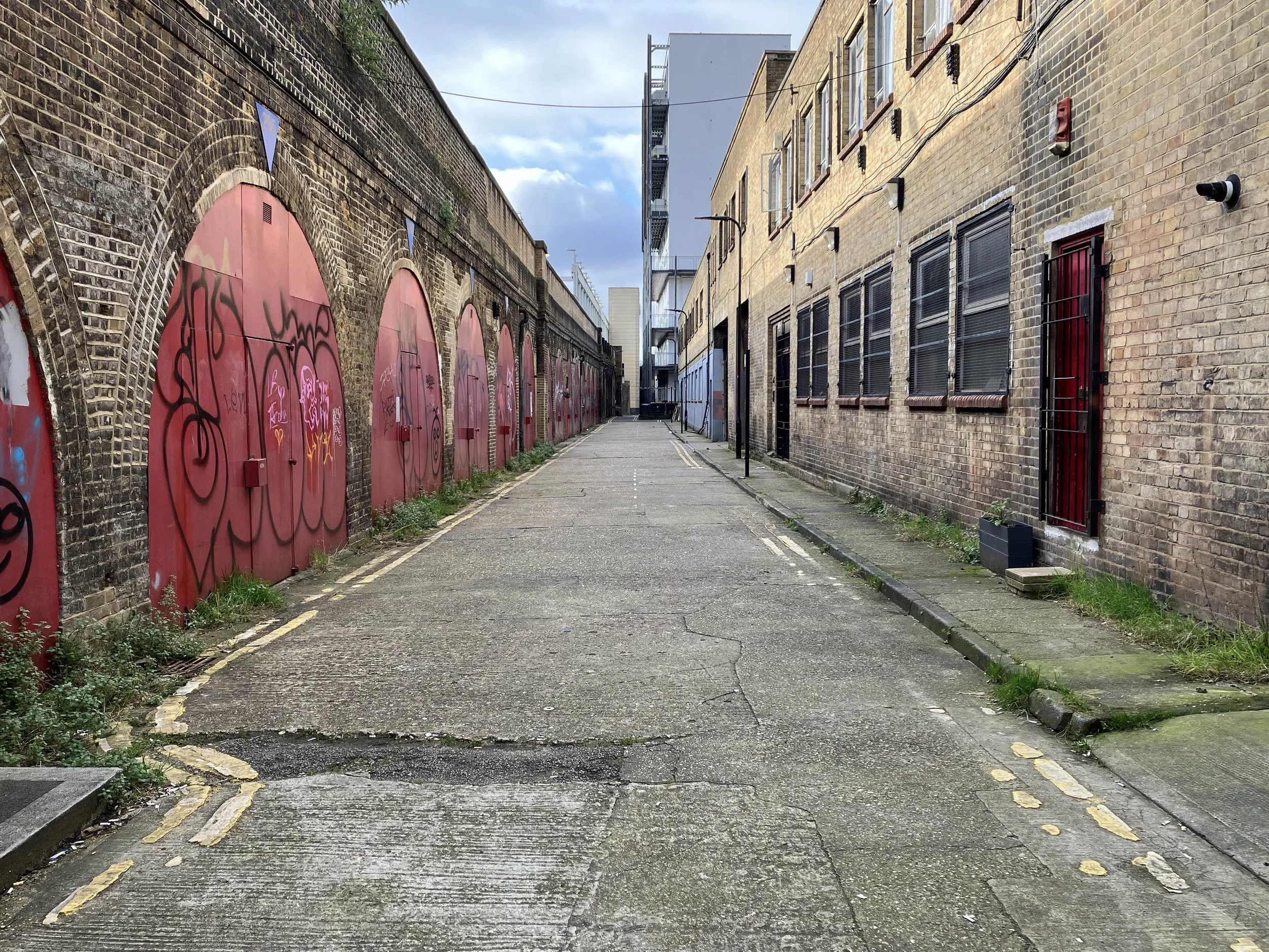An empty alleyway with brick buildings, some with graffiti-covered red doors on the left, and a row of windows with black shutters on the right. The ground is cracked pavement with yellow and white painted lines, and there is a cloudy sky overhead.