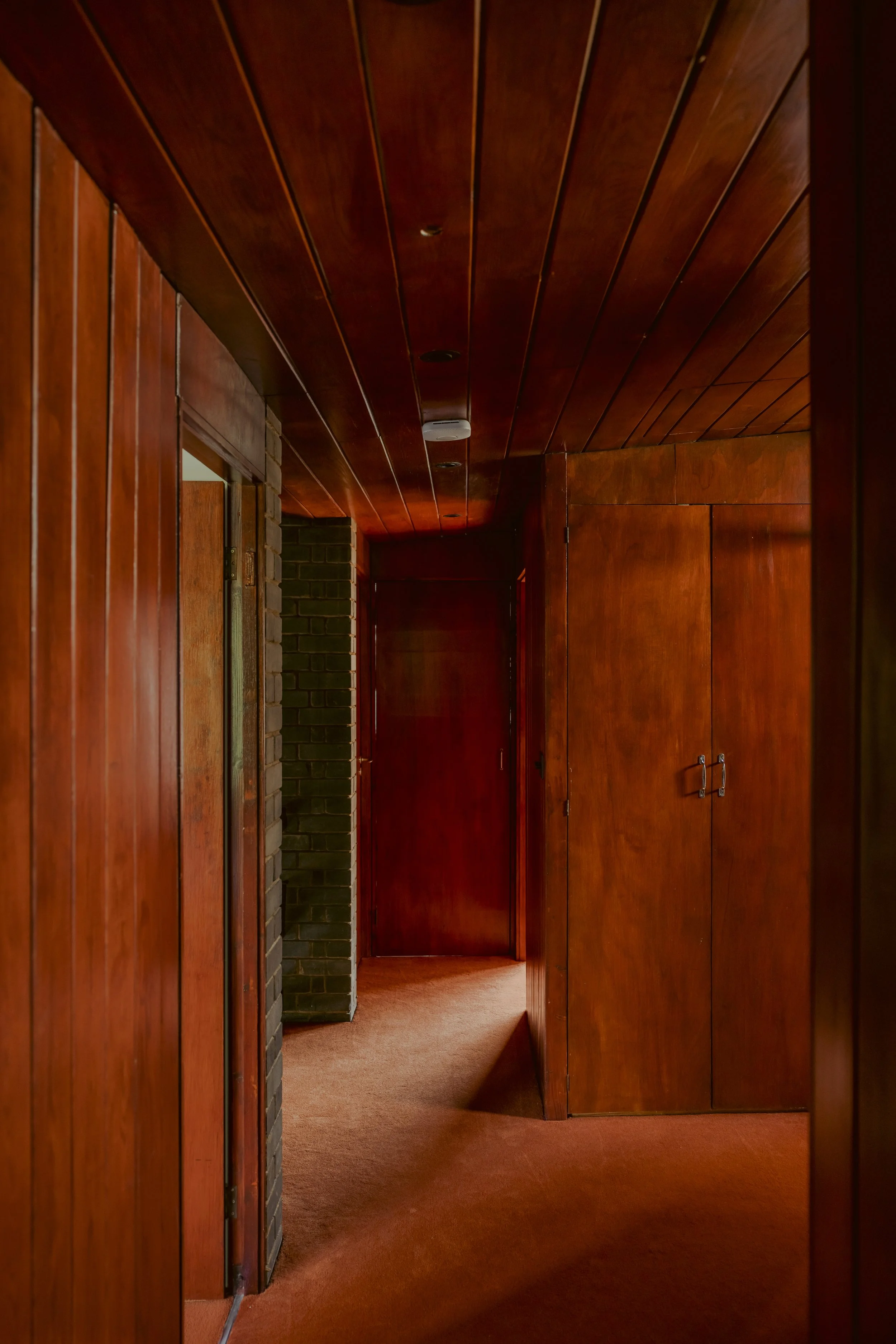 A corridor with wood-paneled walls, wood ceiling, and brown carpeted floor, featuring a brick wall section on the left and several closed wooden doors along the hallway.