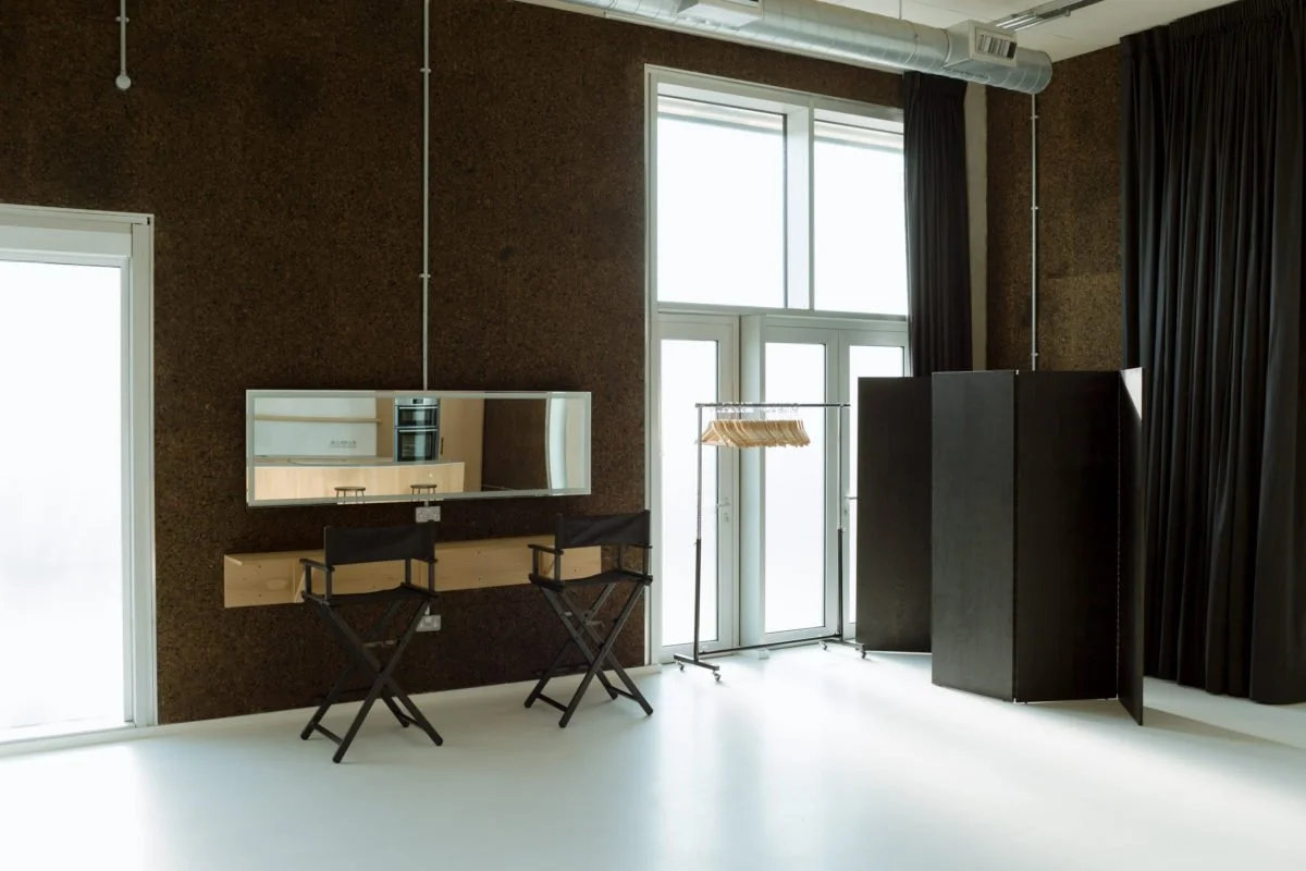 Minimalist interior room with a large window and dark curtains, a wall-mounted mirror, two folding chairs, a clothing rack with hangers, and black folding screens.