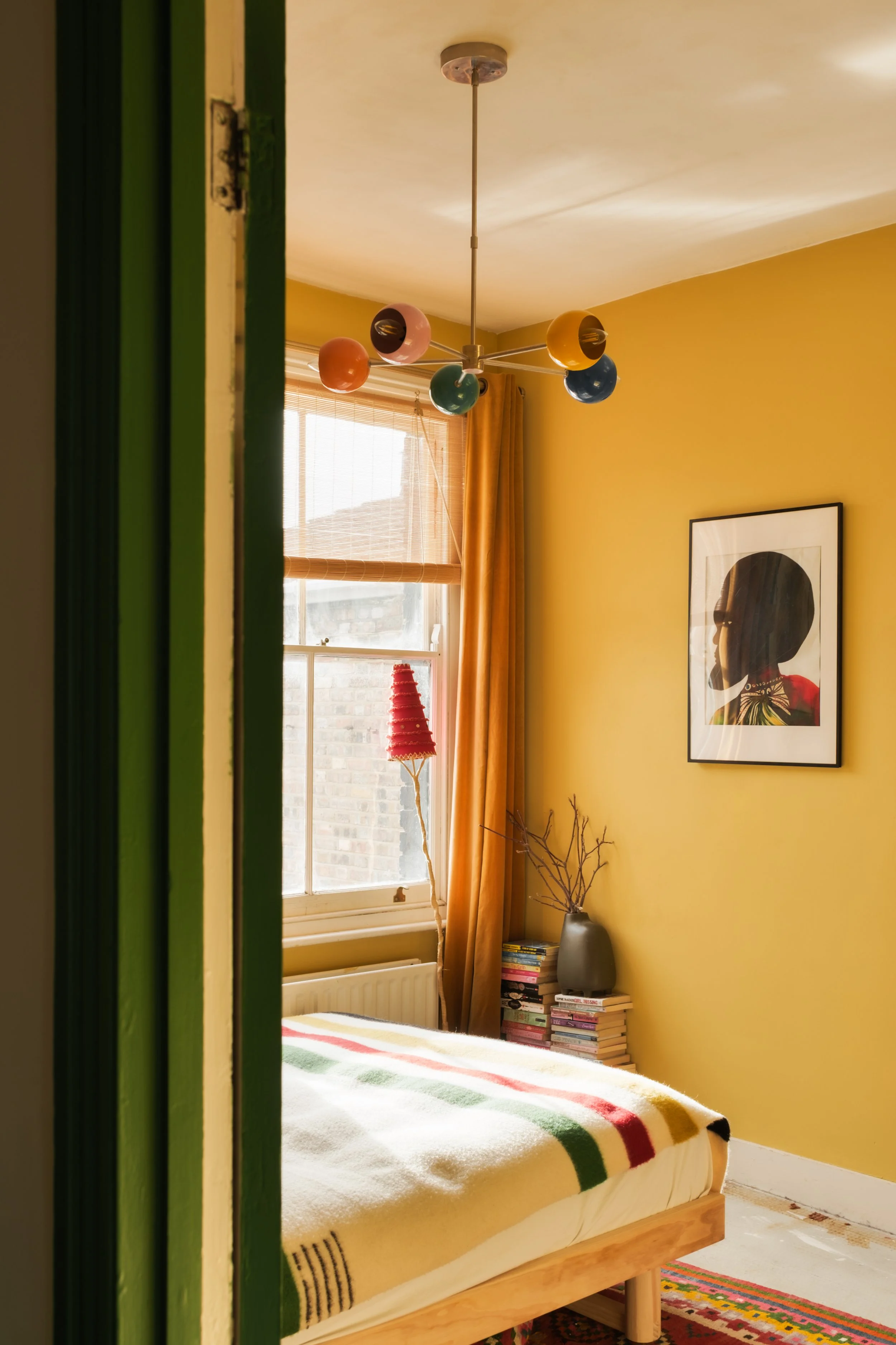 A cozy bedroom with yellow walls and a window with blinds and curtains. The bed has a yellow blanket with red, green, and black stripes. There is a stack of books in a gray vase with dried branches, and a framed silhouette portrait on the wall. A mod