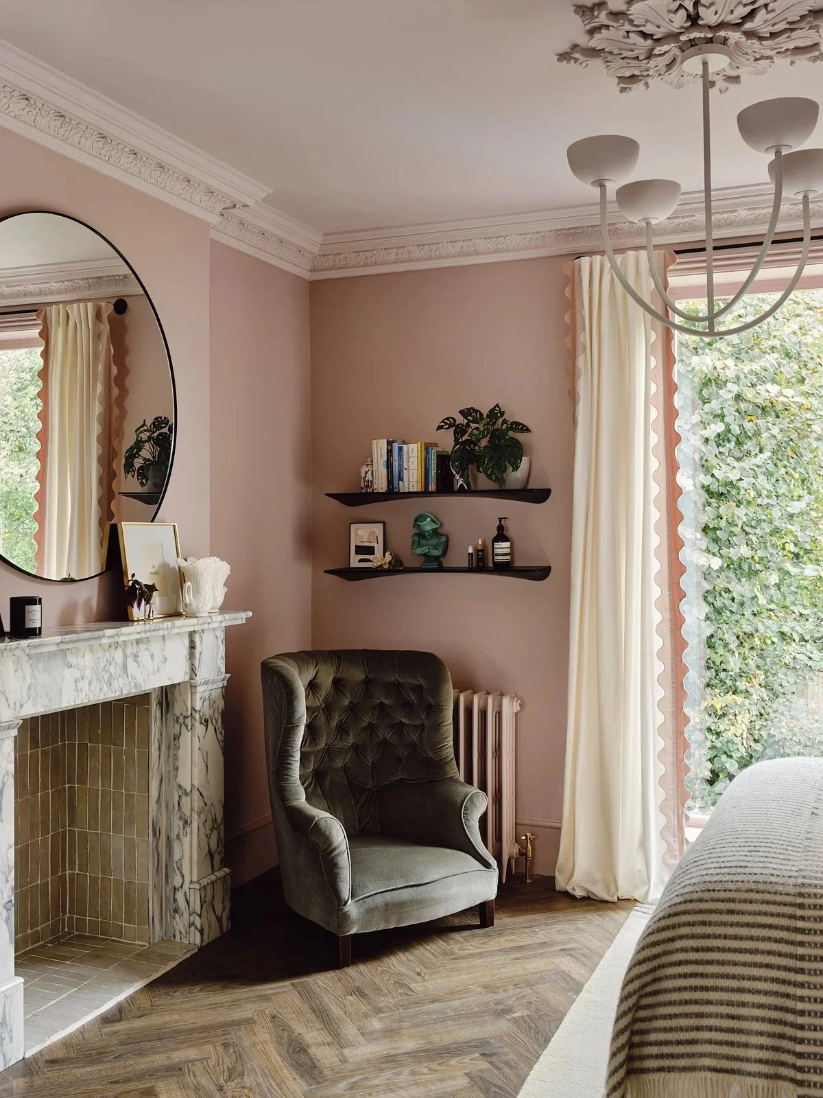 A cozy living room corner with a vintage green armchair, a marble fireplace, and a large window with curtains. Decorative items and books are on wall shelves, and a chandelier hangs from the ceiling.