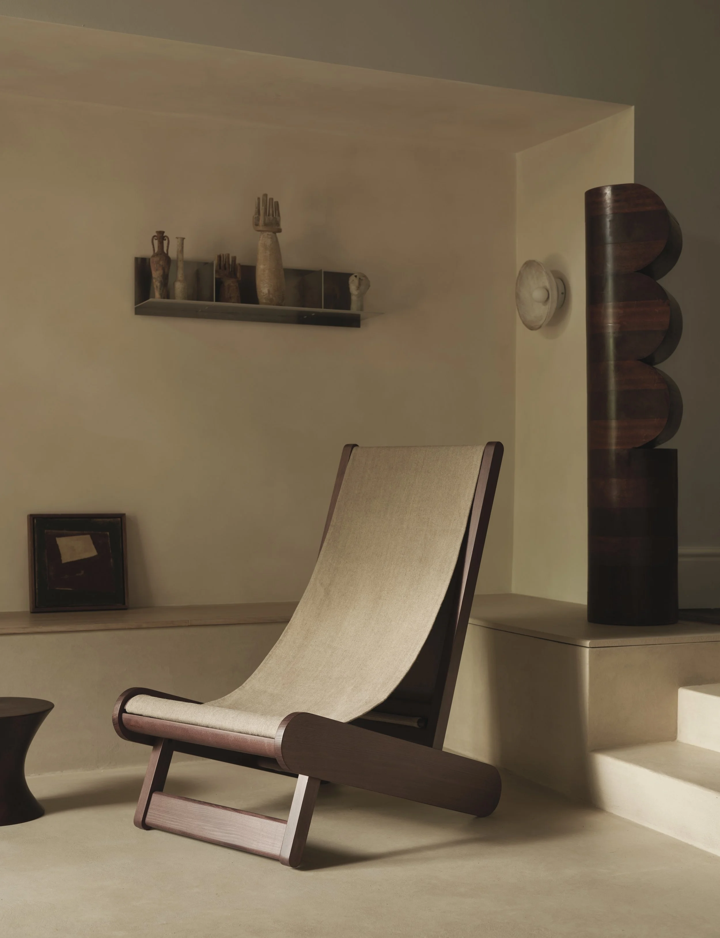 A minimalistic interior with a beige lounge chair, wooden accents, a small framed art piece, ceramic vases on a wall shelf, and a large dark wooden sculpture.