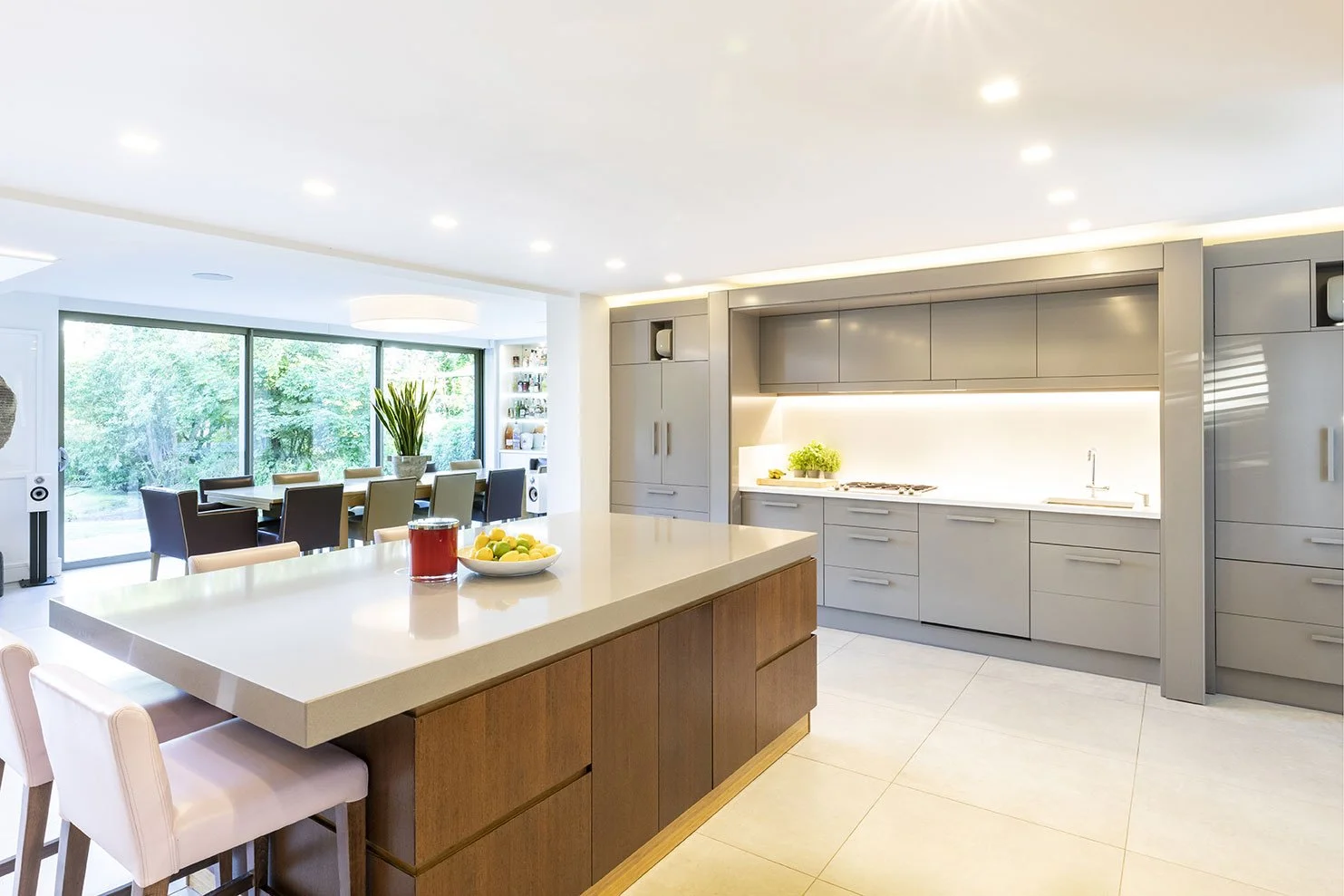 A modern kitchen with a large white island counter, light gray cabinets, and a dining area with a large window and greenery outside.