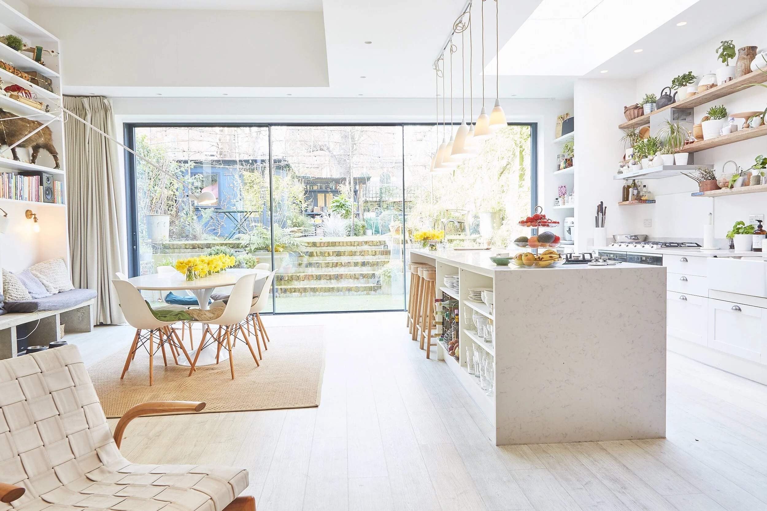Bright open-concept kitchen and dining area with large glass sliding doors leading to a garden. White kitchen island with bar stools, open shelving with plants, and a dining table with white chairs and a yellow flower centerpiece.