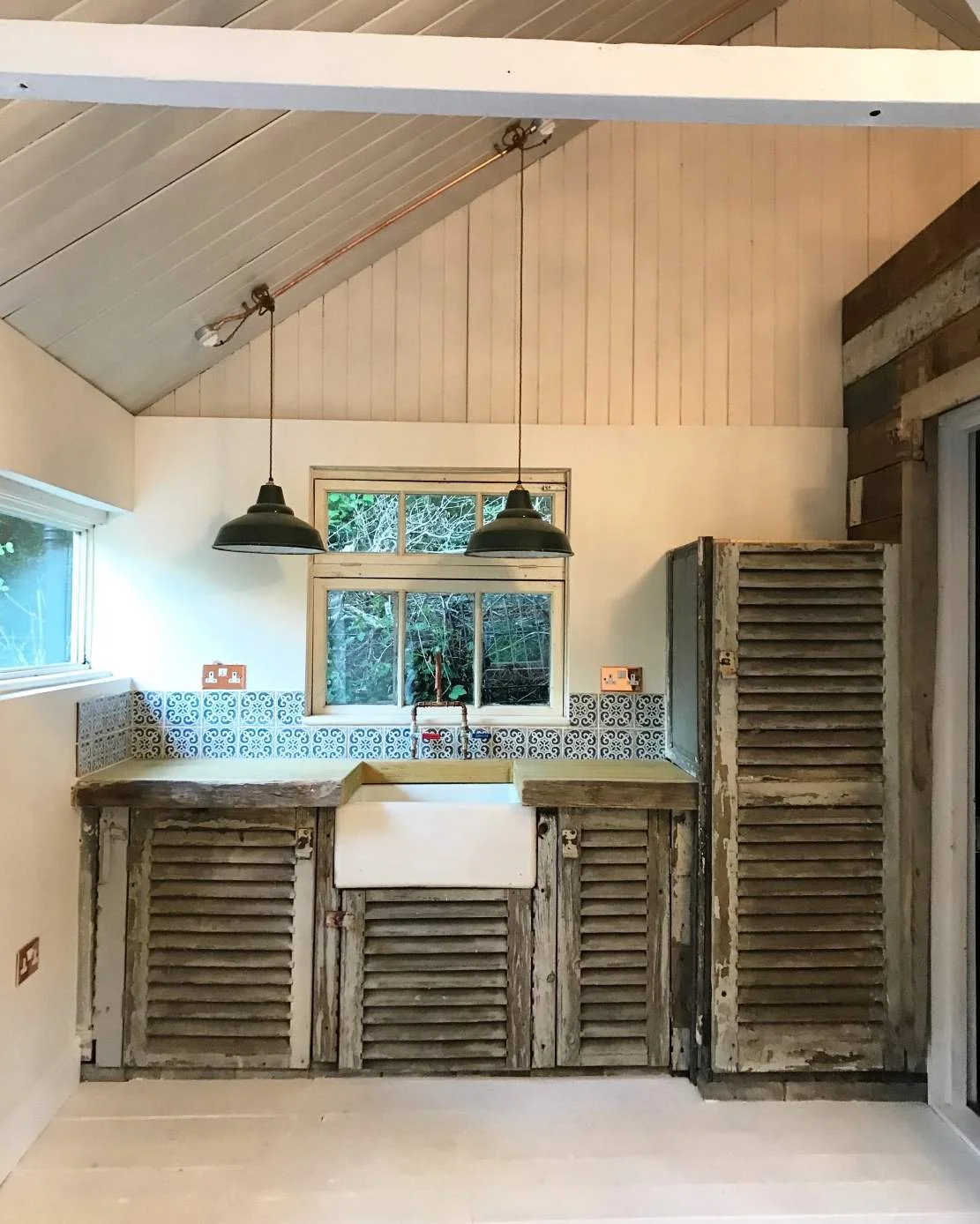 Rustic kitchen with worn wood cabinets, a farmhouse sink, black pendant lights, a window above the sink, and a patterned tile backsplash.
