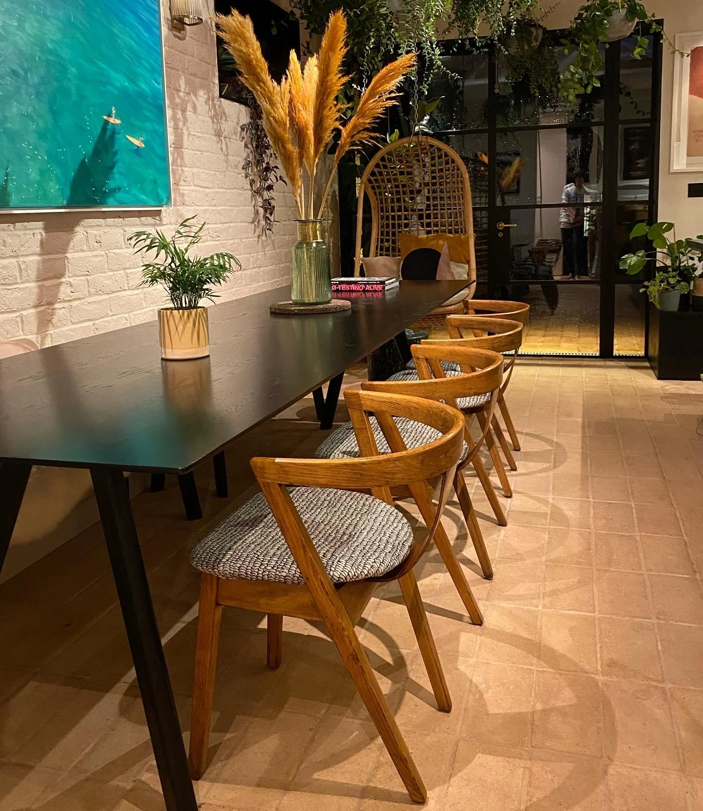 A long dark wooden table with five wooden chairs with upholstered seats, set against a white brick wall decorated with a large painting of a blue sea scene. On the table are two vases with plants and pampas grass, and a pink book. In the background, 