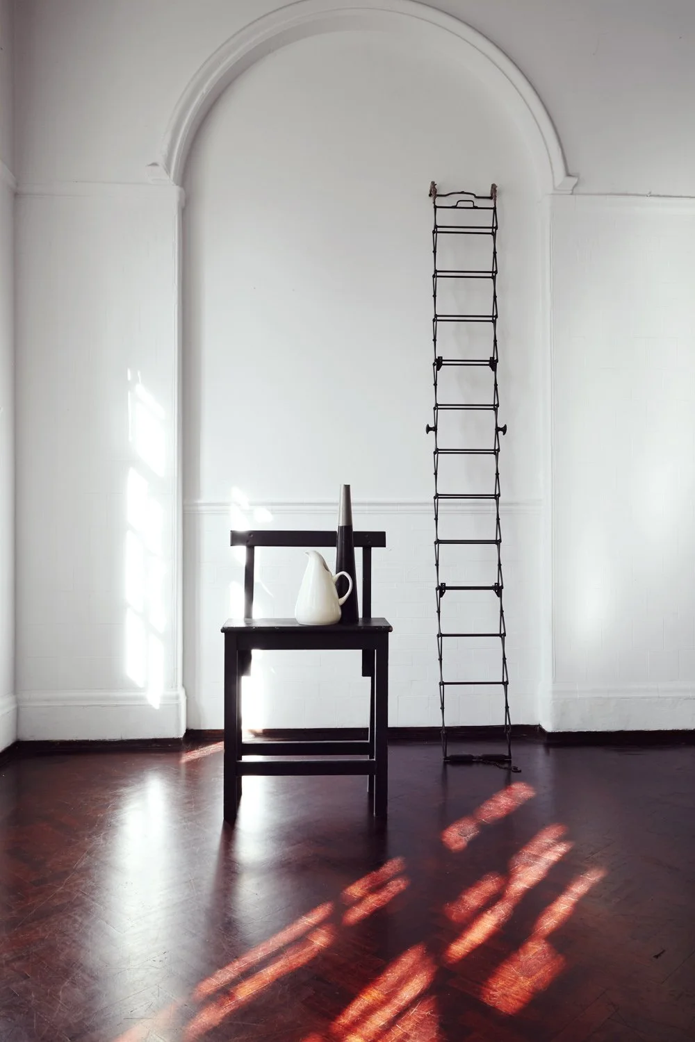 A minimalist interior with white walls, dark wooden floor, a black chair with a white pitcher and a black cone on top, and a tall, narrow, black foldable ladder leaning against a wall with sunlight casting patterned shadows on the floor.