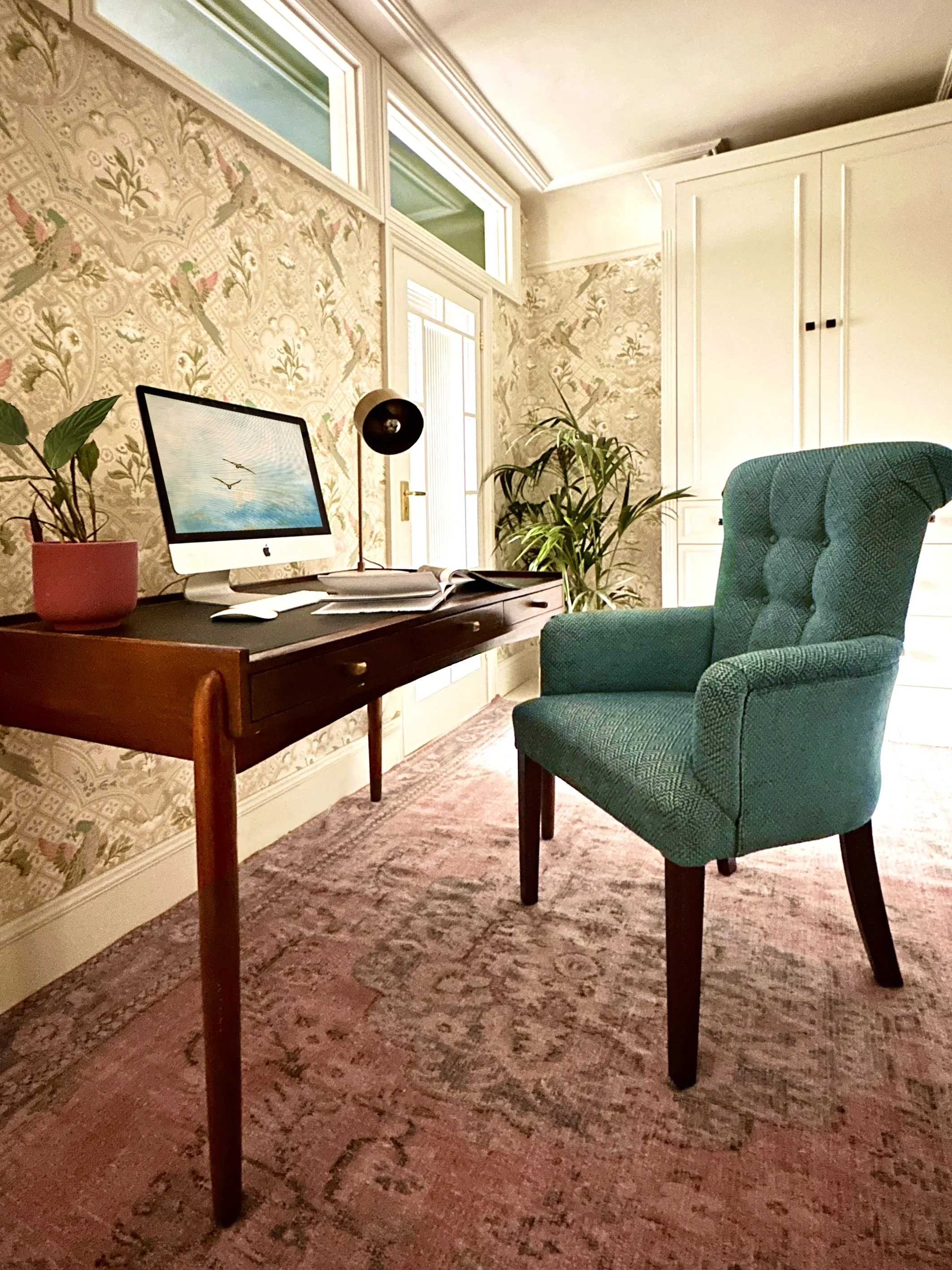 A cozy home office with a vintage wooden desk, an iMac computer, a black desk lamp, a pink potted plant, papers and notebooks, a teal upholstered armchair, green plants, wallpaper with a bird and floral pattern, a pinkish rug, and a white cabinet in 