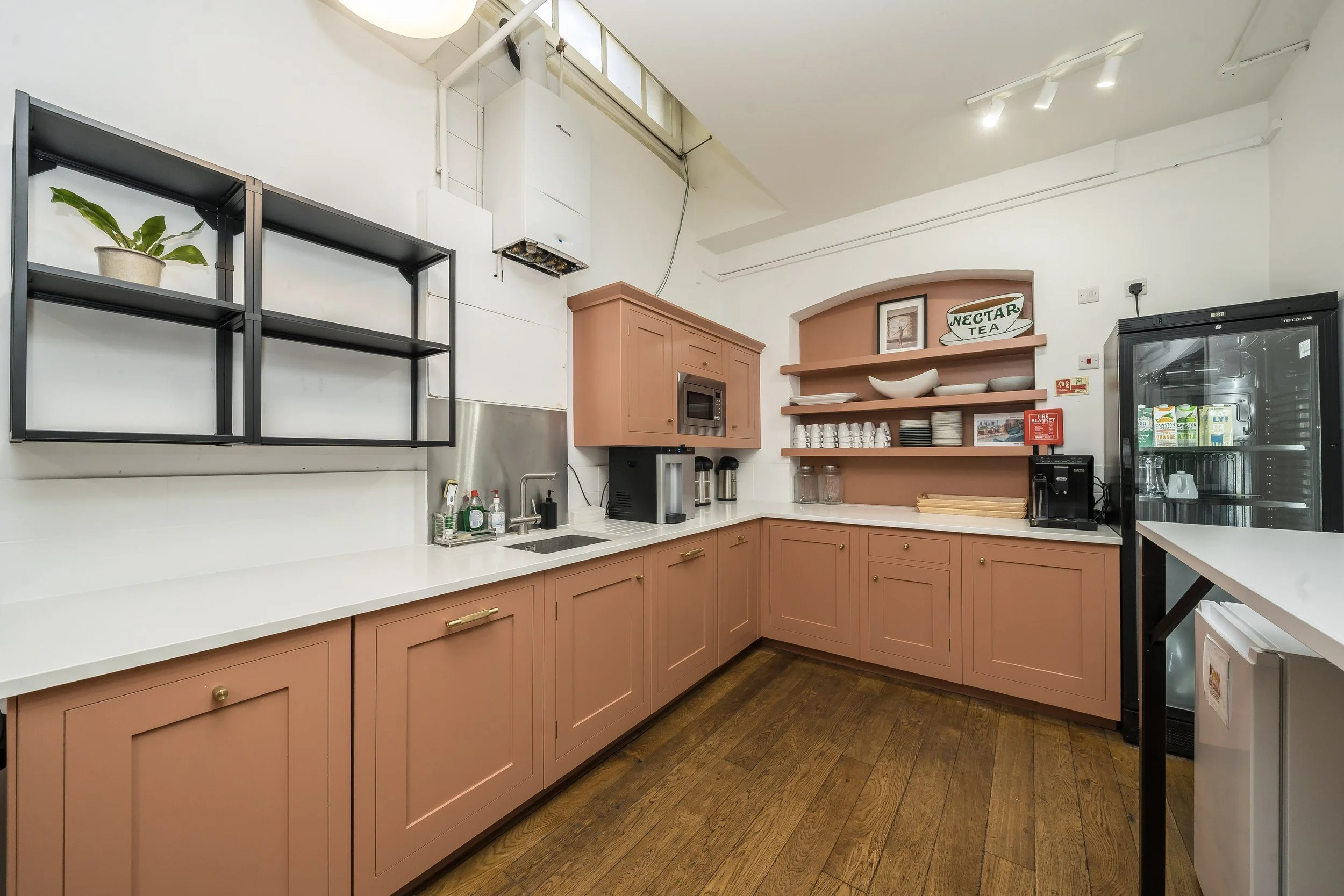 Kitchen with pink cabinets, white countertops, open shelves, a microwave, coffee machines, a refrigerator, and a plant on black wall-mounted shelf.