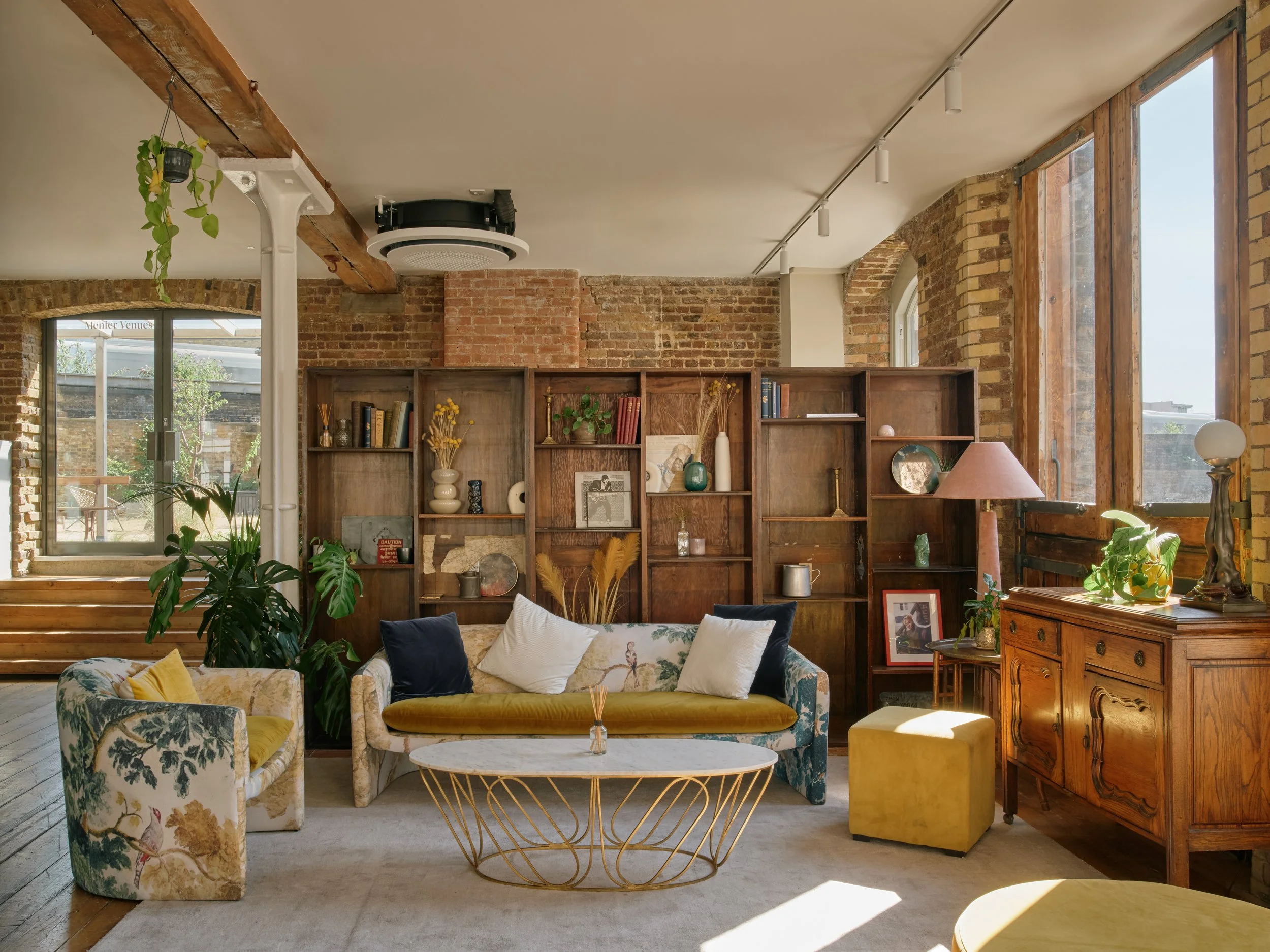 Living room with exposed brick walls, wooden furniture, large windows, and eclectic decor including a floral-patterned armchair, a velvet sofa with pillows, a gold wire coffee table, a wooden sideboard, and shelves with decorative items and books.